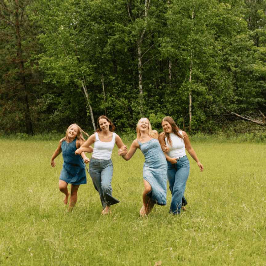 Four happy women running through a lush green meadow surrounded by trees on a sunny day.