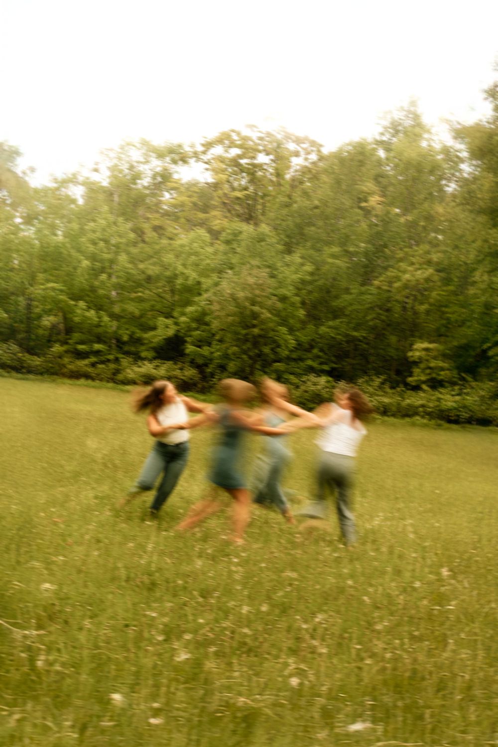 Blurred motion photo of friends holding hands and spinning in a circle on a green meadow with trees in background.