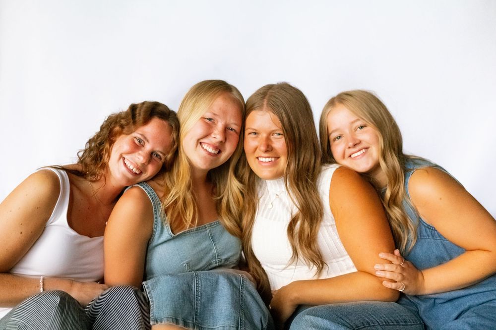Four smiling young women in denim overalls pose together against a white background in a cheerful group portrait.