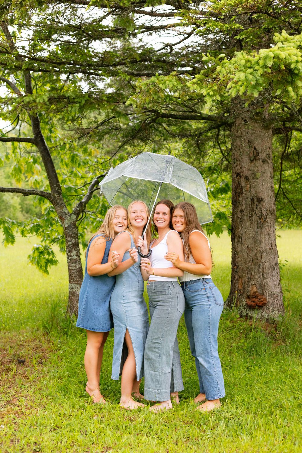 Four young women smiling under a clear umbrella outdoors among green trees in denim outfits.
