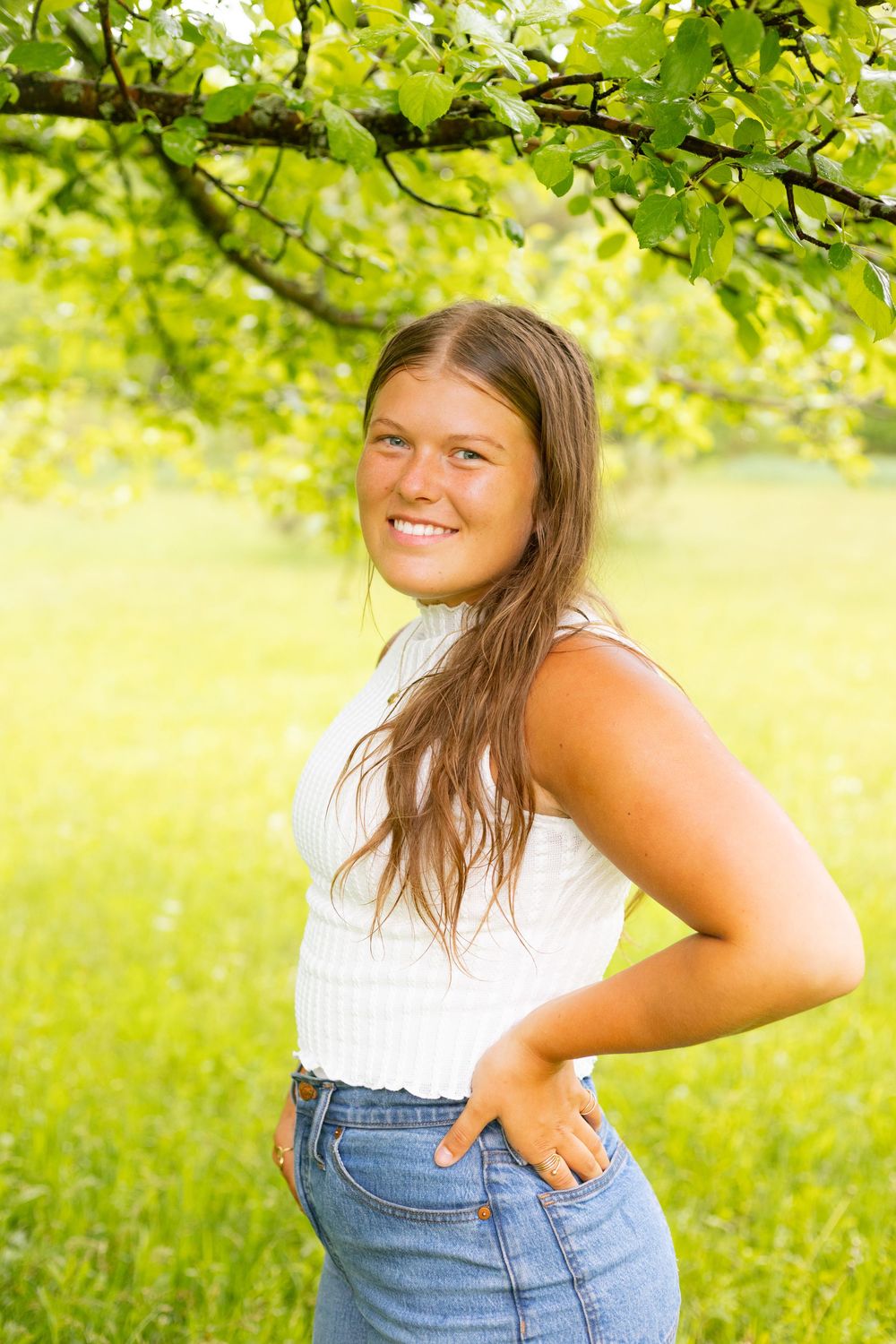 Smiling young woman in white top and jeans posing outdoors under green tree branches on a sunny day.