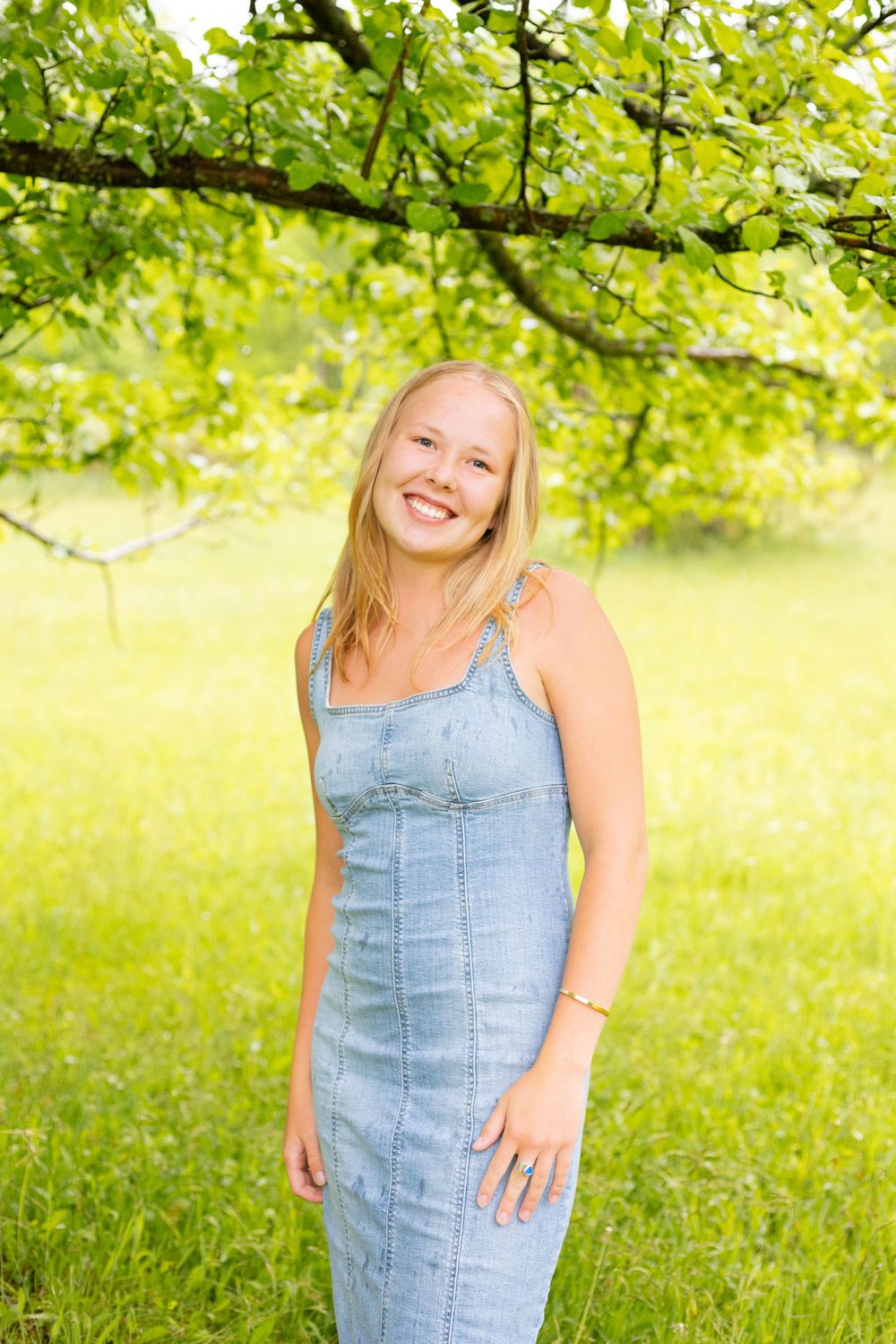 Smiling blonde woman in a denim dress standing outdoors under a tree with lush green foliage in the background.