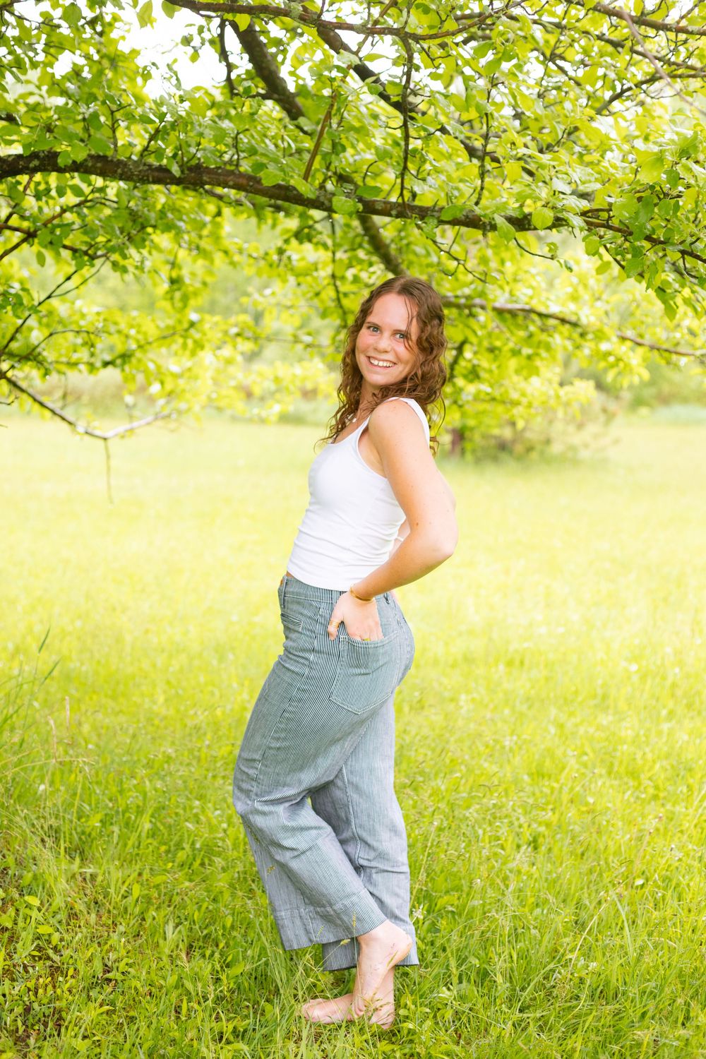 Smiling young woman in white tank top and jeans standing under green tree branches in sunny park.