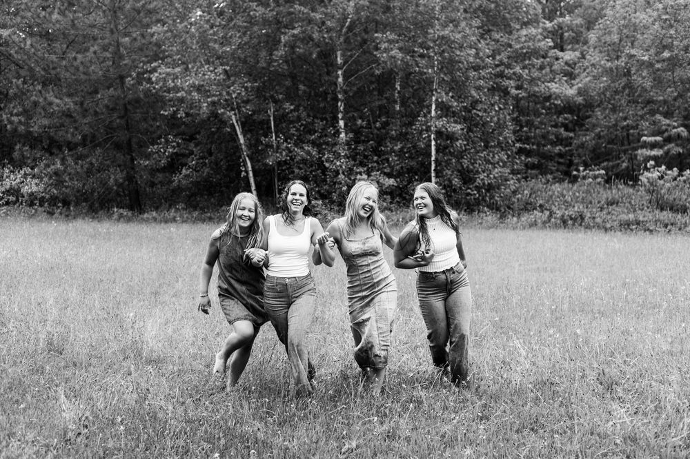 Four women laughing and running together through a grassy field with trees in the background, black and white photo.