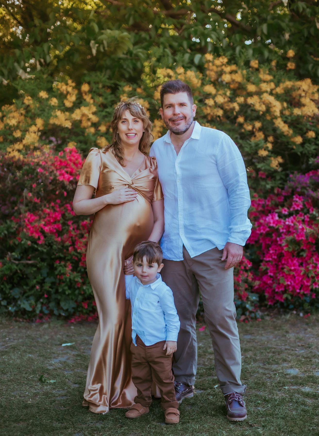 Pregnant woman in gold gown poses with husband and toddler in front of colourful flowering  backdrop. During a family session in London