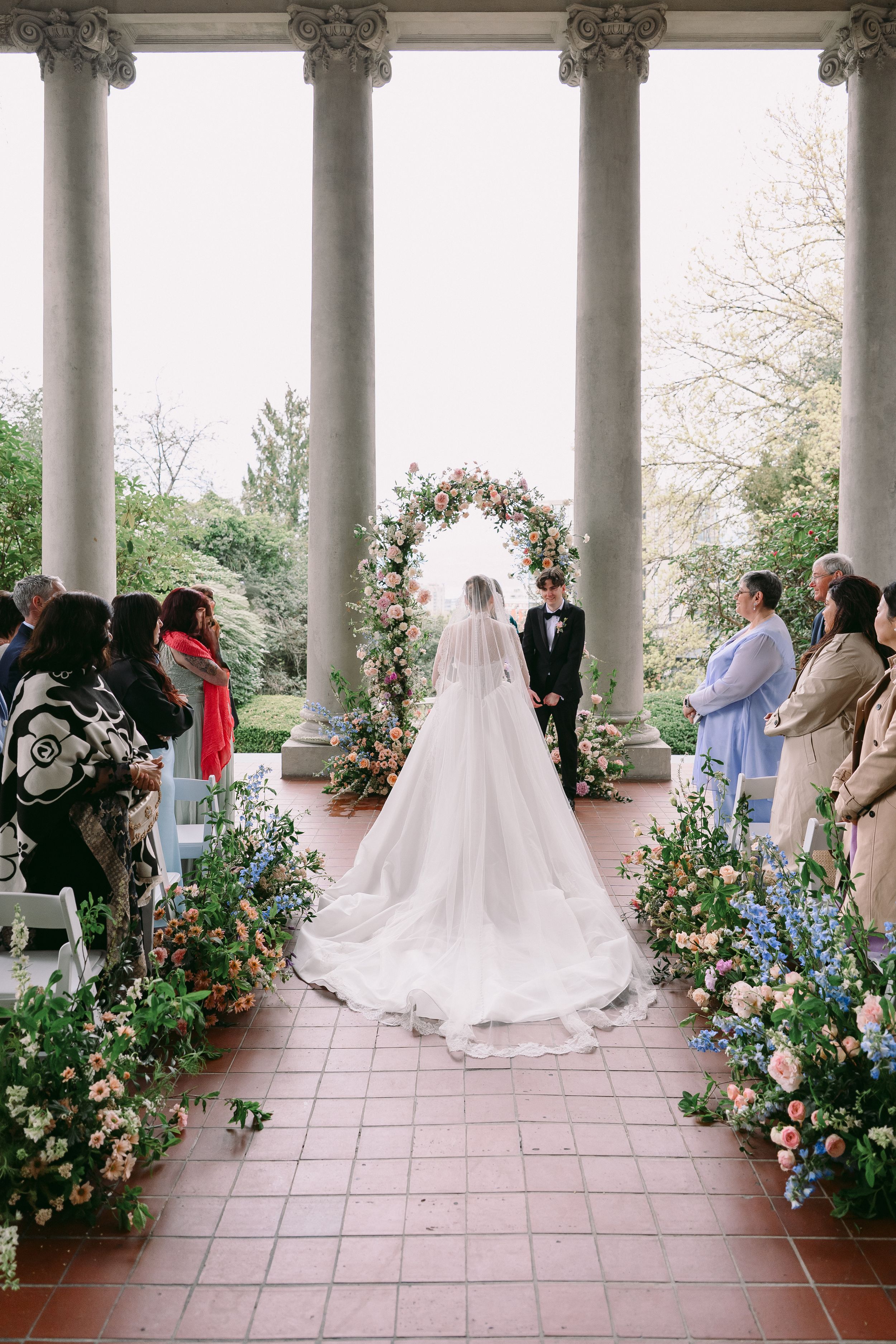 Bride and groom at floral arch altar in columned garden pavilion, aisle lined with colorful blooms and guests standing.