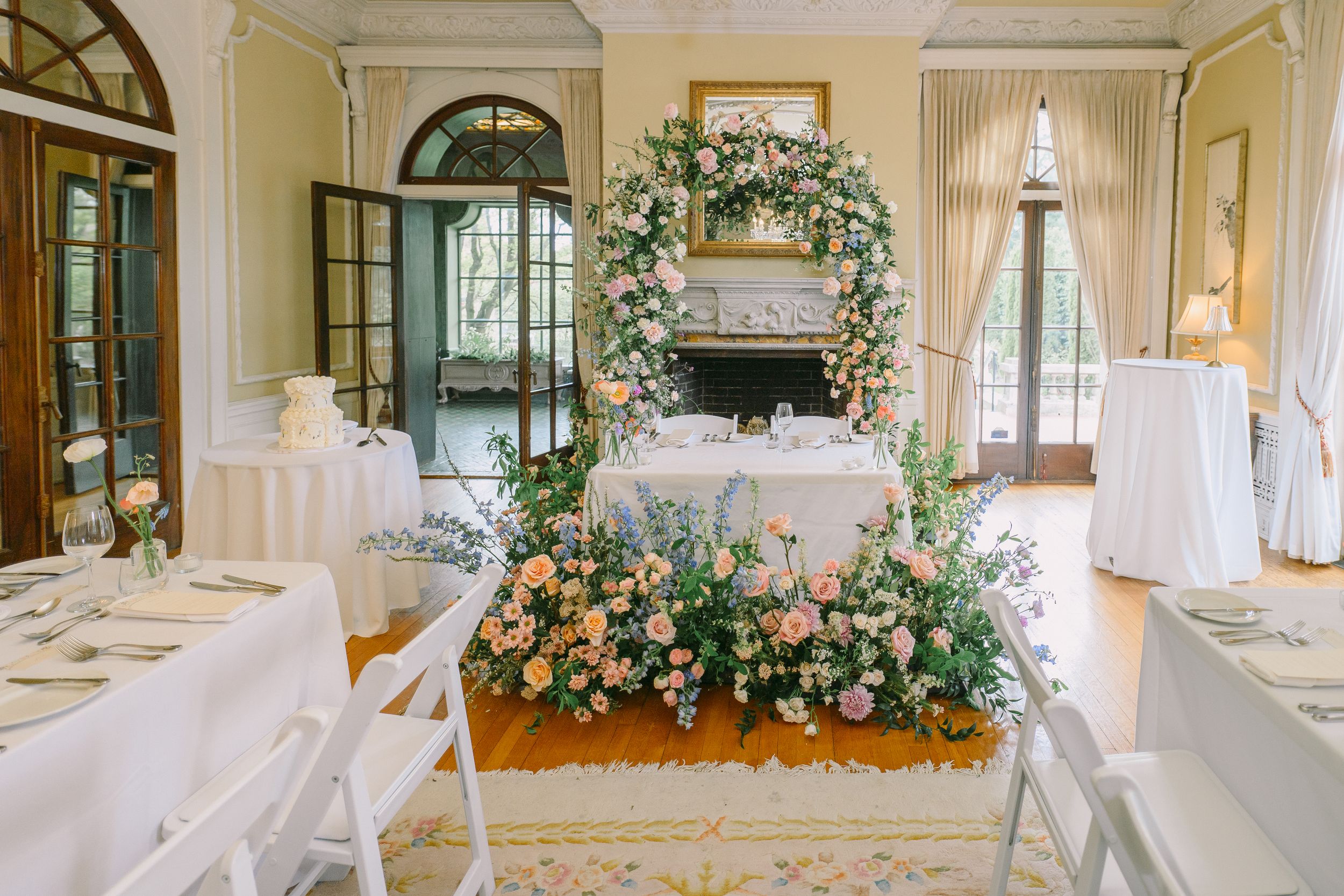 Elegant wedding reception room with floral arch, peach and pink blooms on ceremony table, white chairs, and ornate fireplace backdrop.