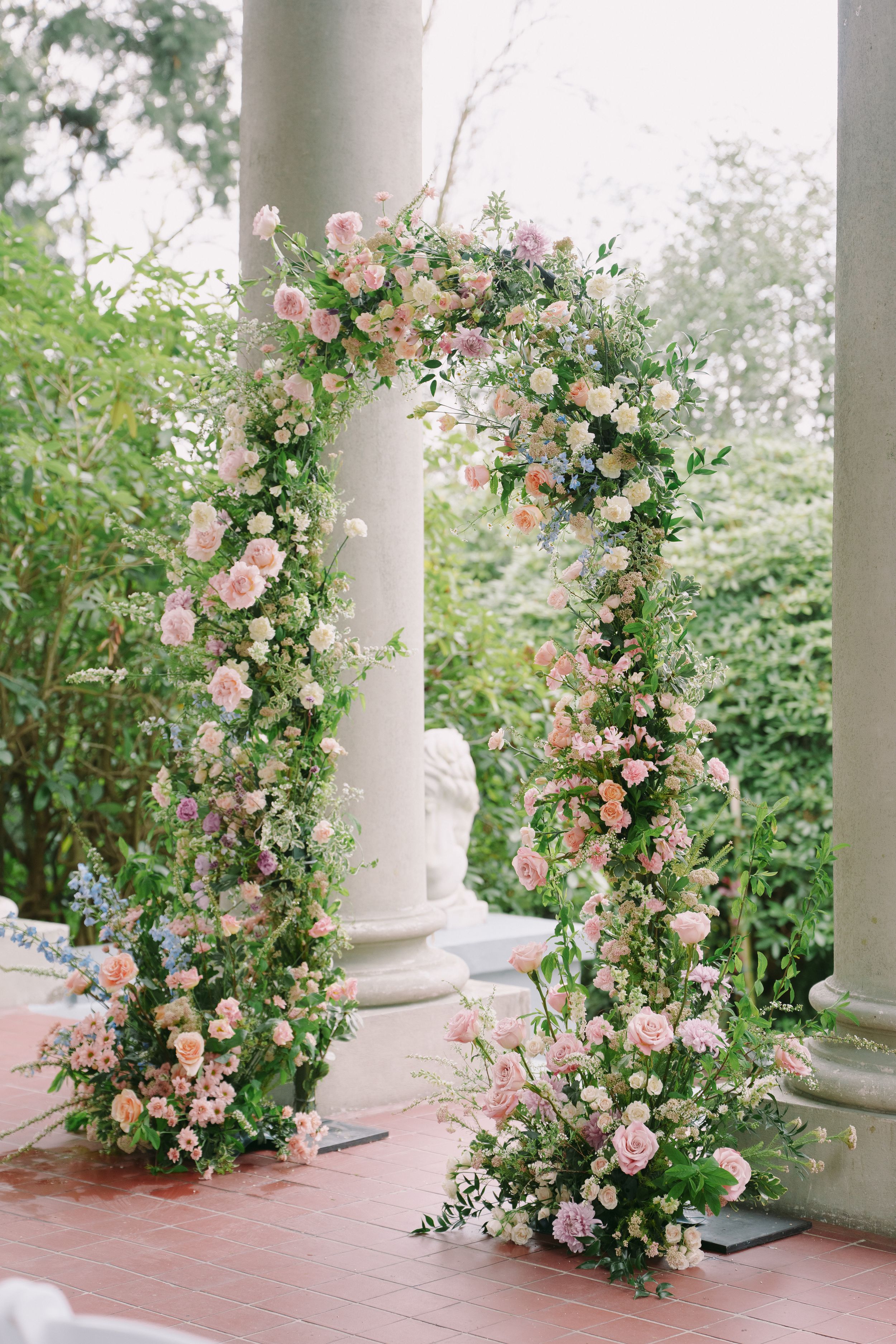 Elegant floral arch with pink roses and greenery framing a garden wedding ceremony near white columns.