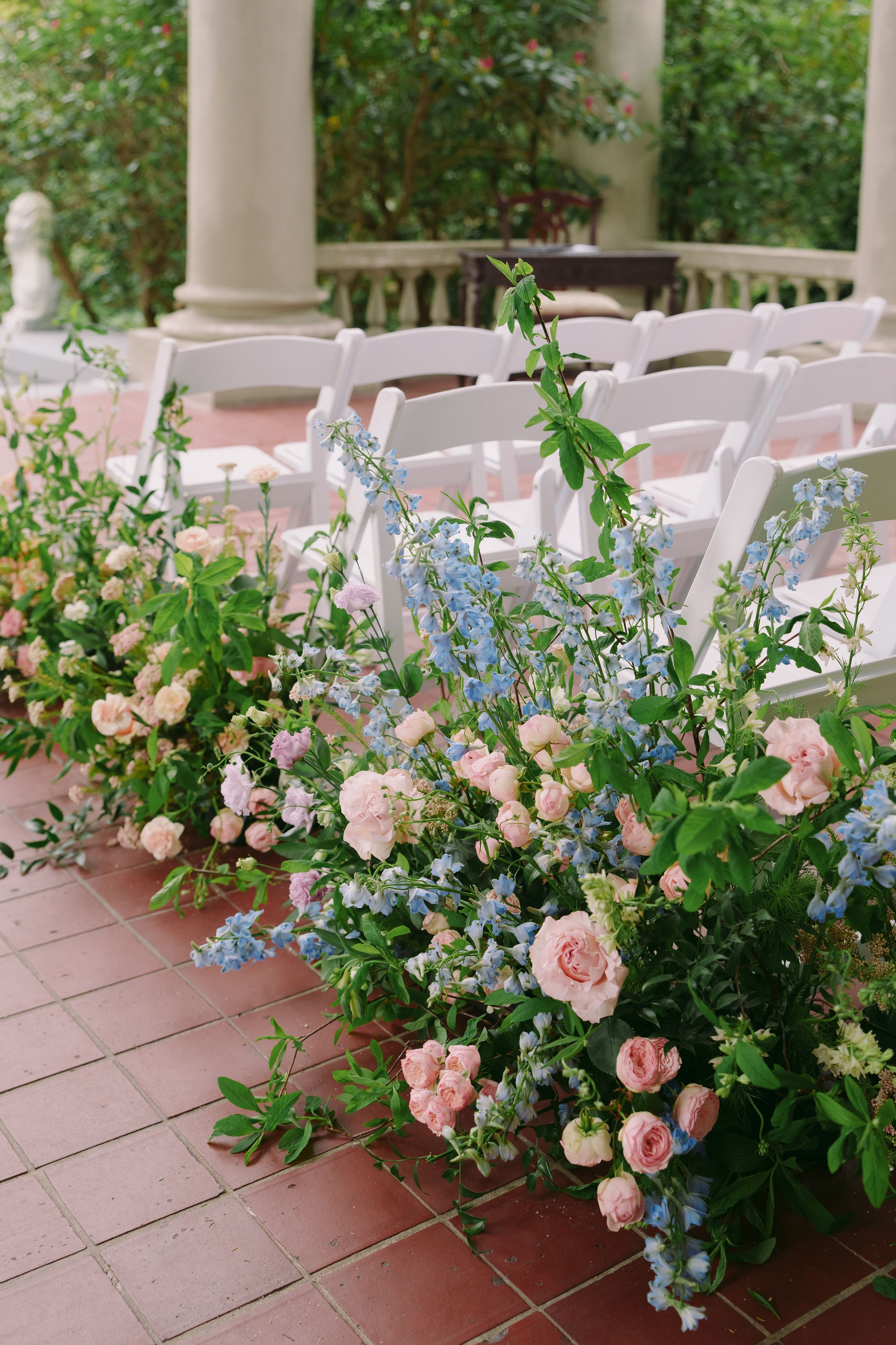 Lush pink roses and blue delphiniums line a brick aisle with white chairs at an elegant outdoor wedding ceremony.