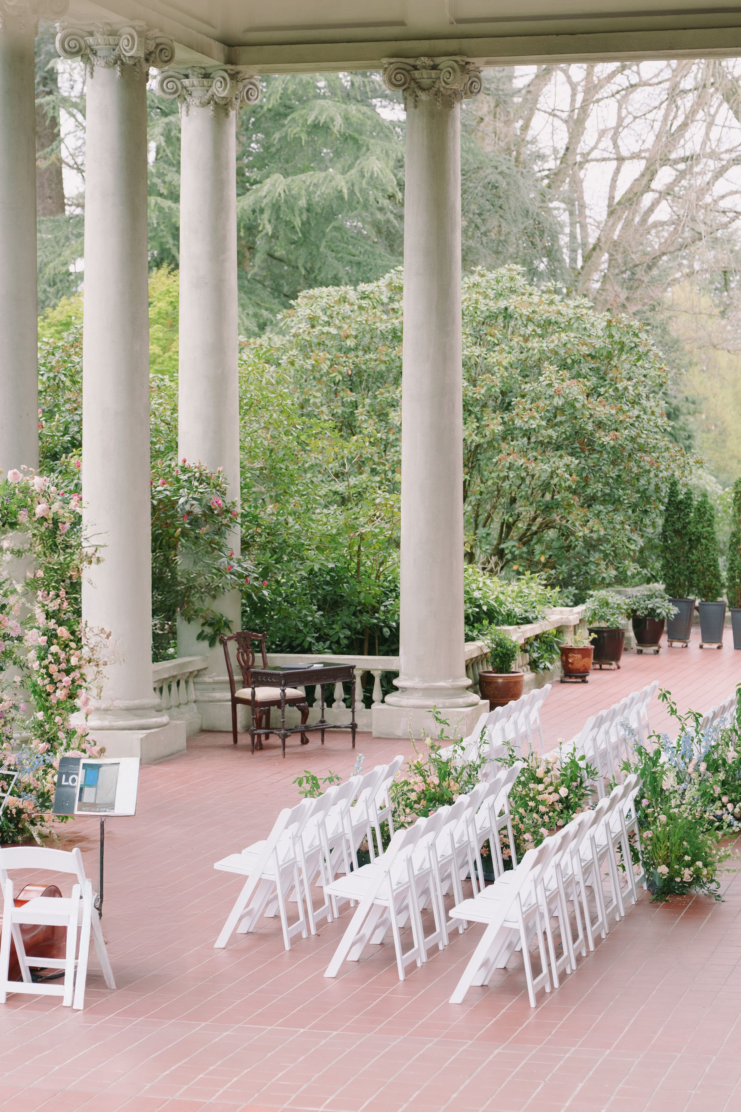 Elegant outdoor wedding ceremony setup with white folding chairs, tall stone columns, and lush garden greenery on a brick terrace.