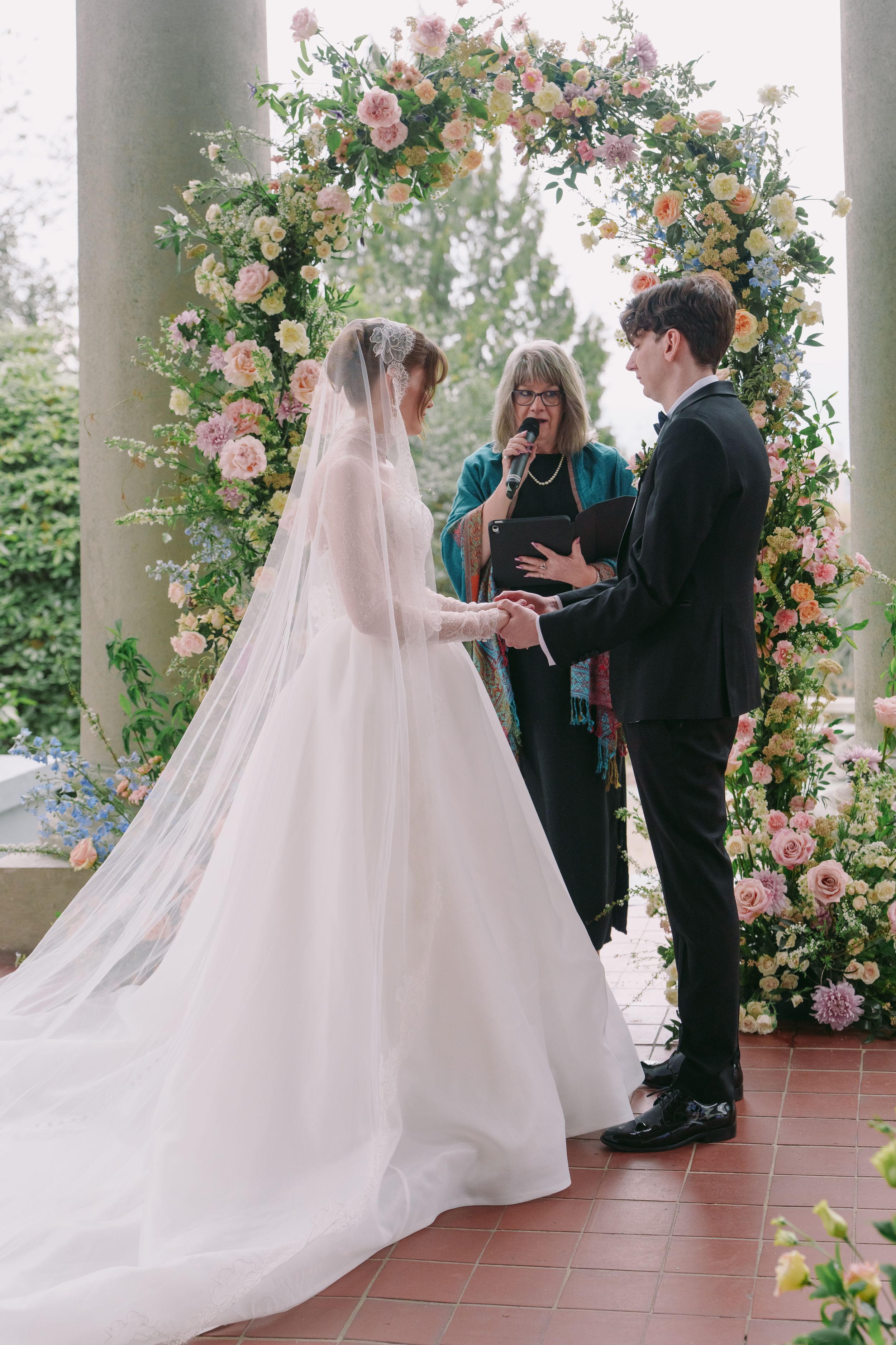 Bride and groom exchange vows under a floral arch with pink and white blooms, officiated by a woman in teal.