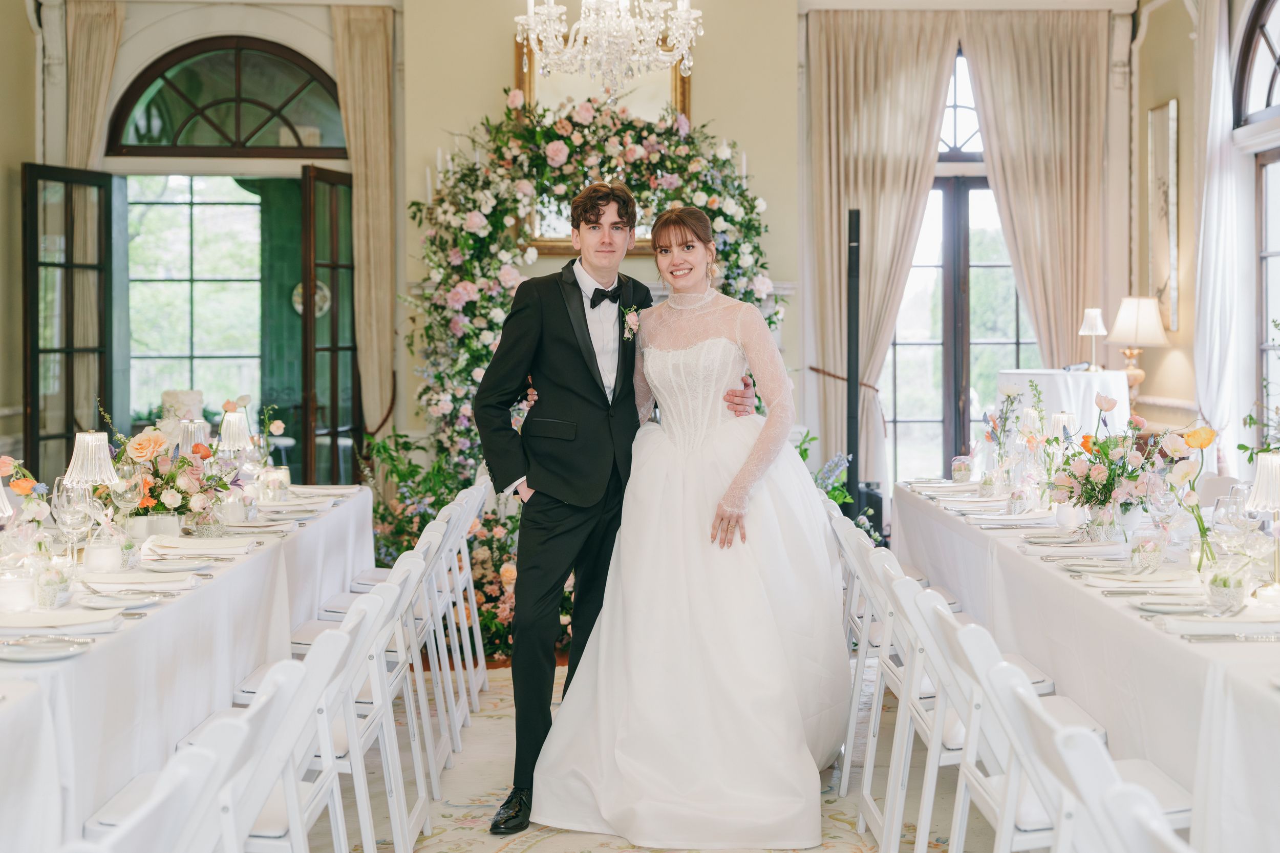 Elegant wedding couple poses in a floral-decorated reception hall with white chairs and tables.