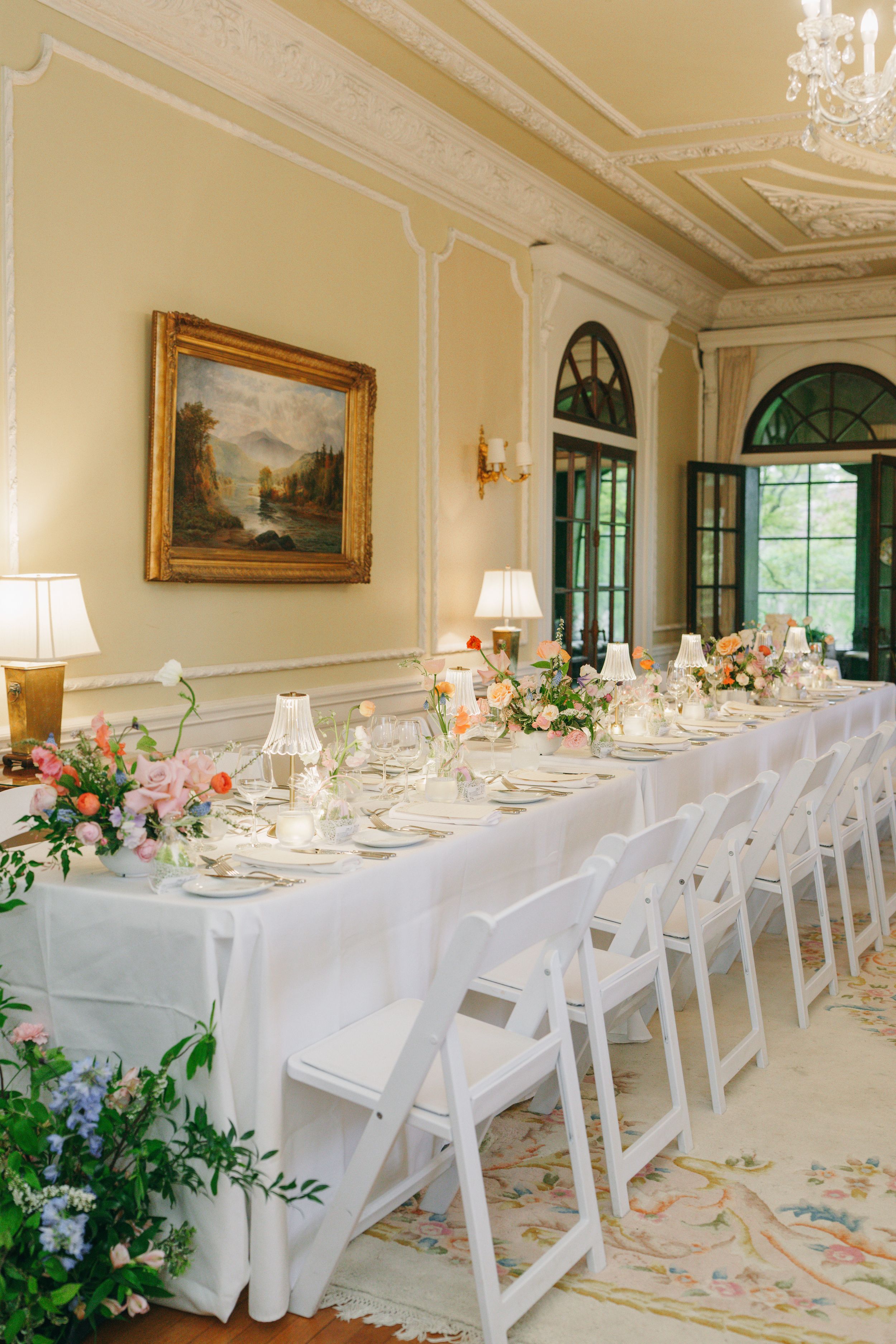 Elegant wedding reception table with white linens, floral centerpieces, and classic white chairs in a grand ballroom.