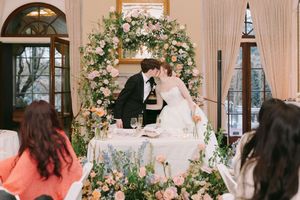 Bride and groom share a kiss at floral arch sweetheart table during elegant indoor wedding reception.