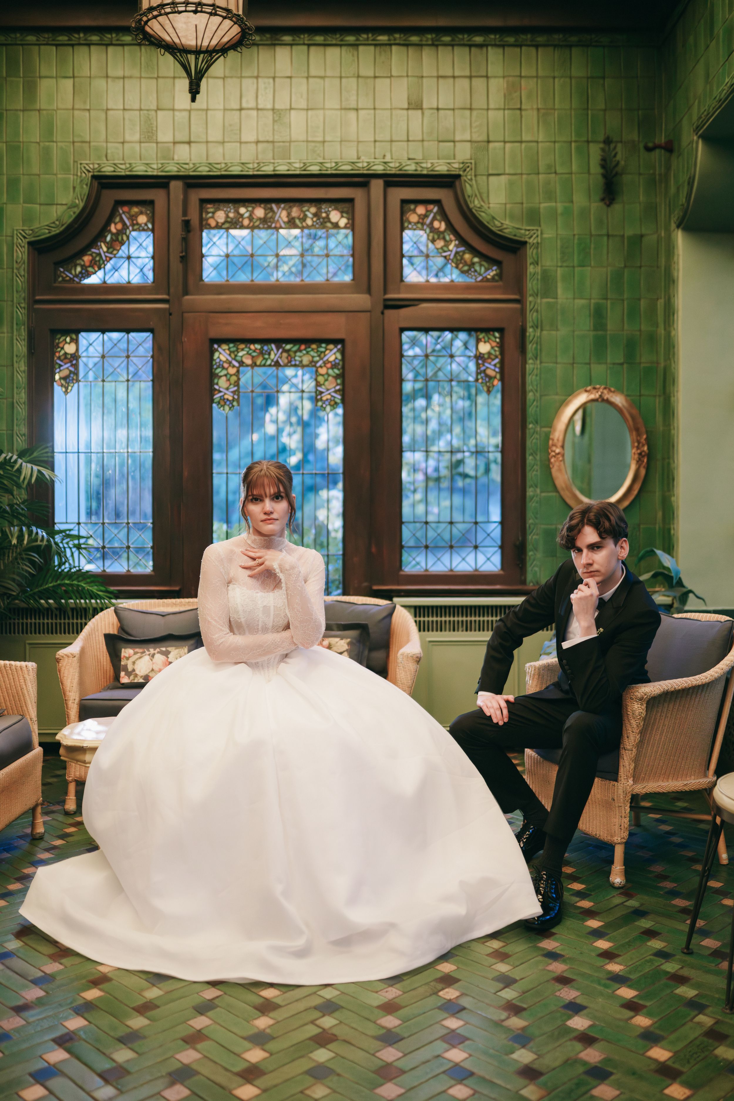 Bride in white ballgown and groom in black suit seated in ornate green-tiled room with stained glass windows.