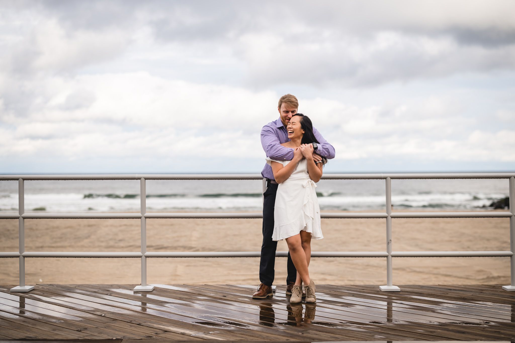 Couple embracing romantically on a beachfront boardwalk with ocean waves and cloudy sky backdrop.