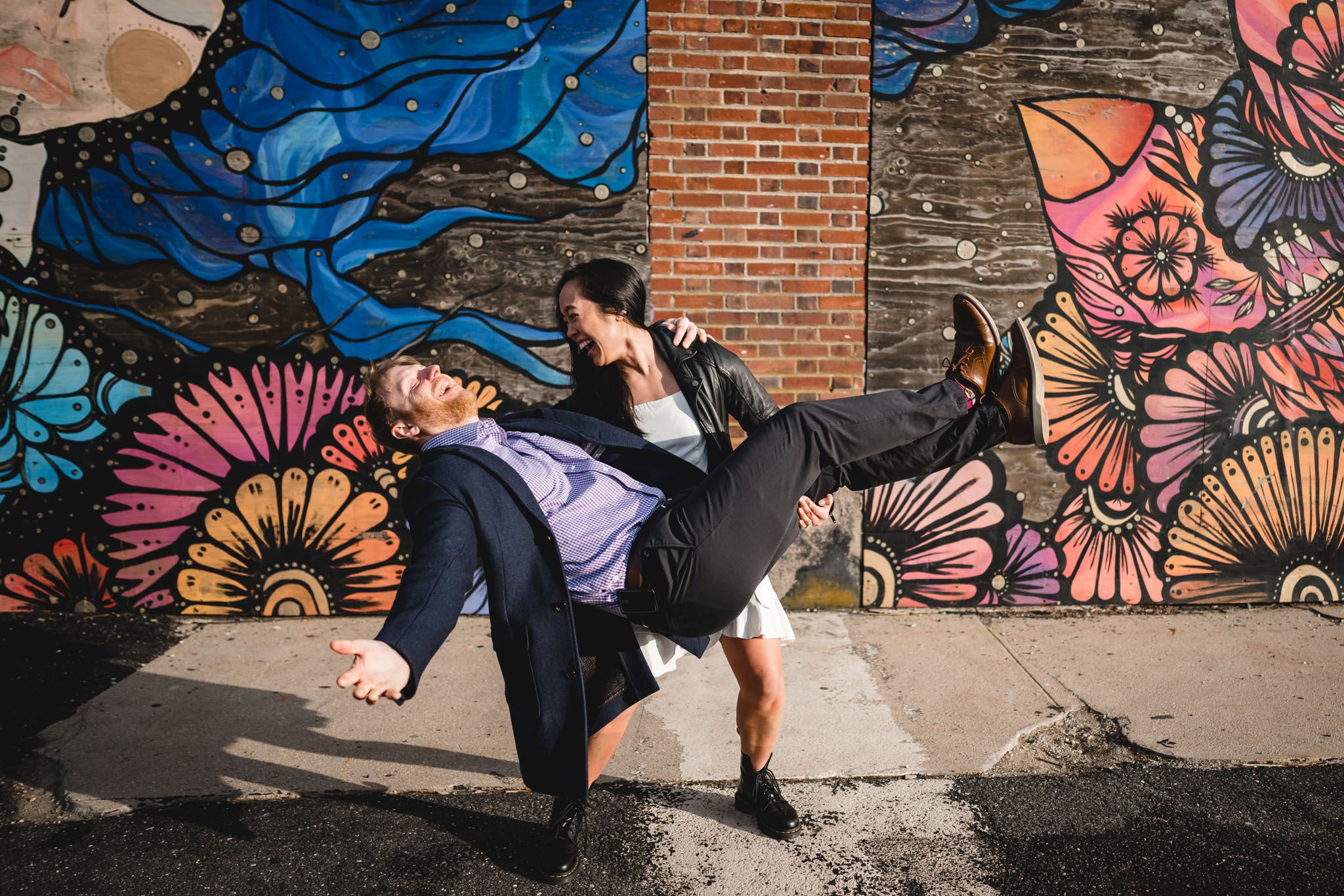 Two people dancing playfully in front of a vibrant colorful floral mural on a brick wall outdoors.