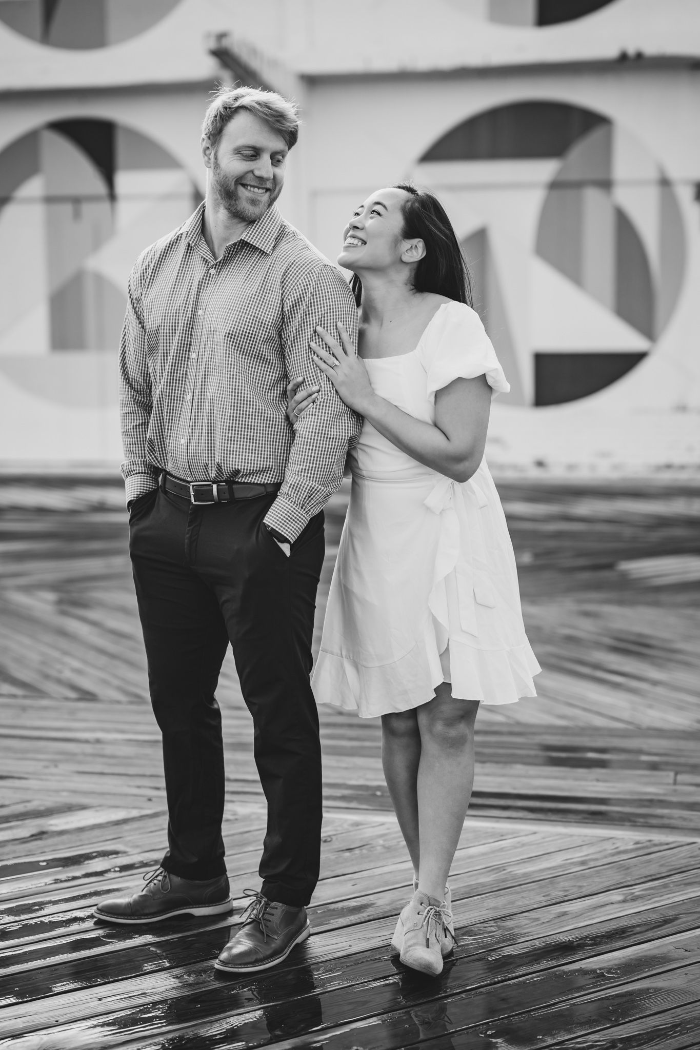 Smiling couple embracing on a wet boardwalk, black and white engagement photo with geometric mural backdrop.