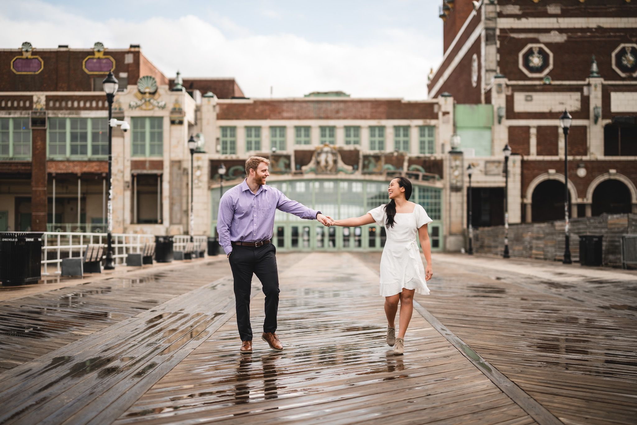 Couple holding hands on a wet boardwalk with historic buildings in the background on a rainy day.