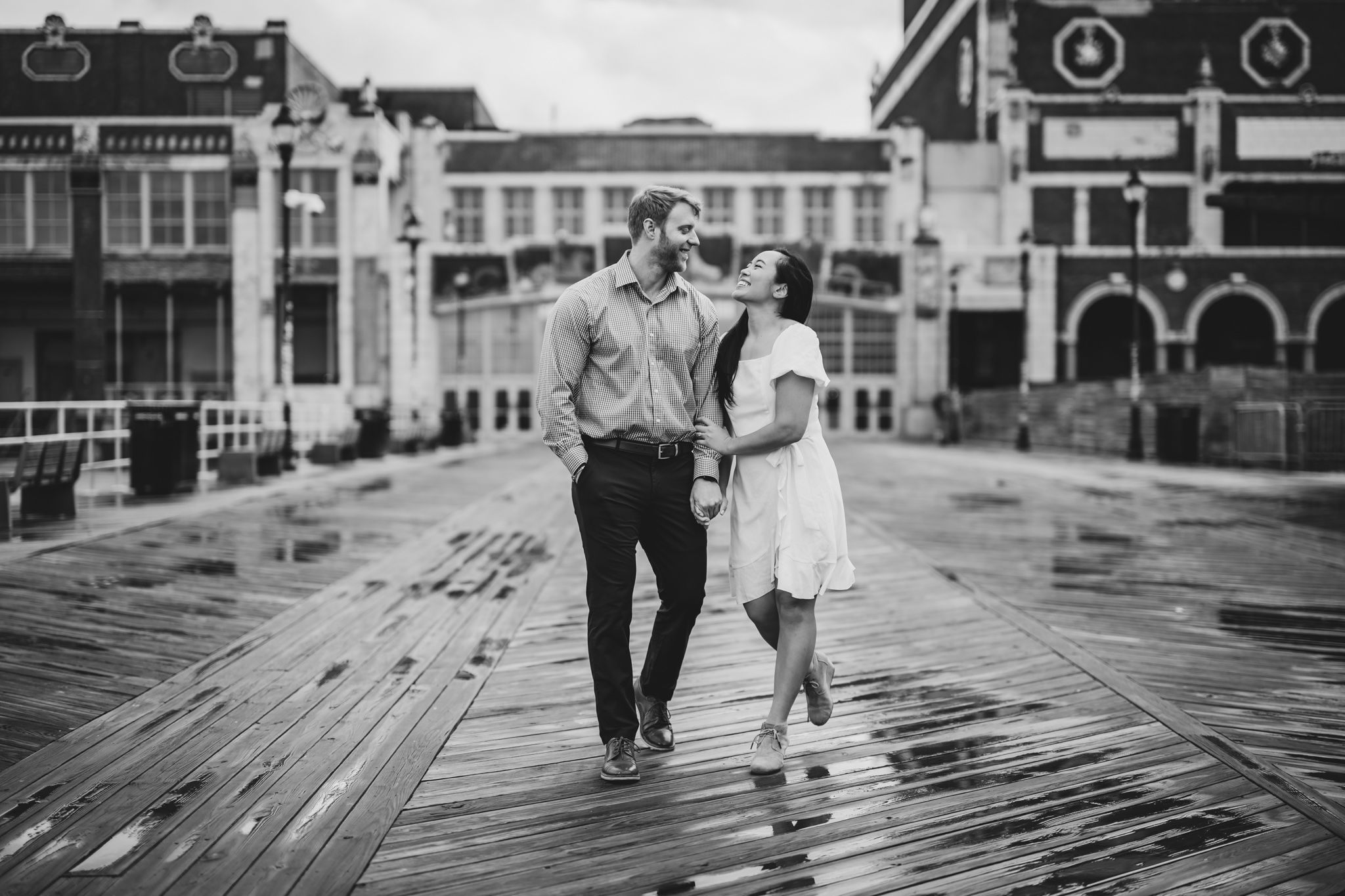 Couple walking hand-in-hand on a wet boardwalk in black and white engagement photo.