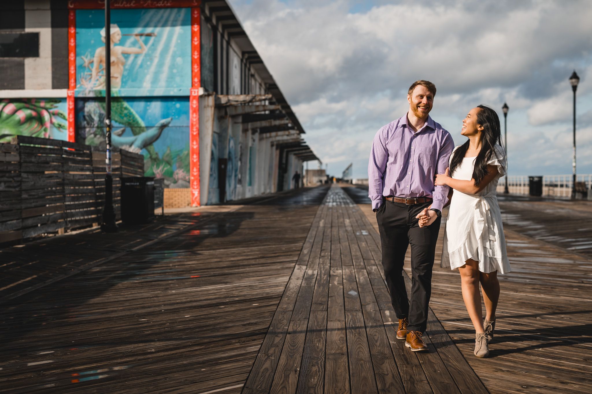 Happy couple walking hand in hand on a sunny boardwalk with colorful murals during engagement session.