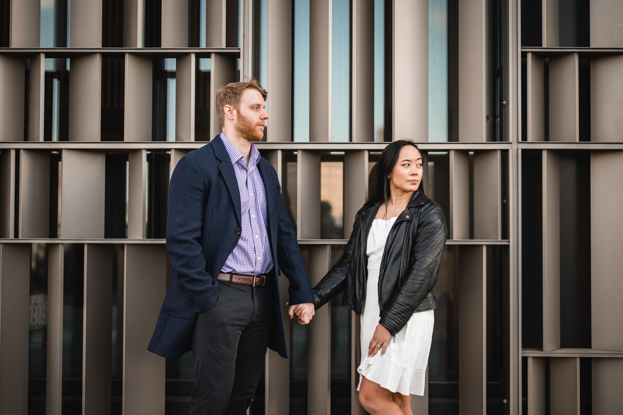 A stylish couple holds hands in front of a modern building, man in blue blazer, woman in leather jacket and white dress.