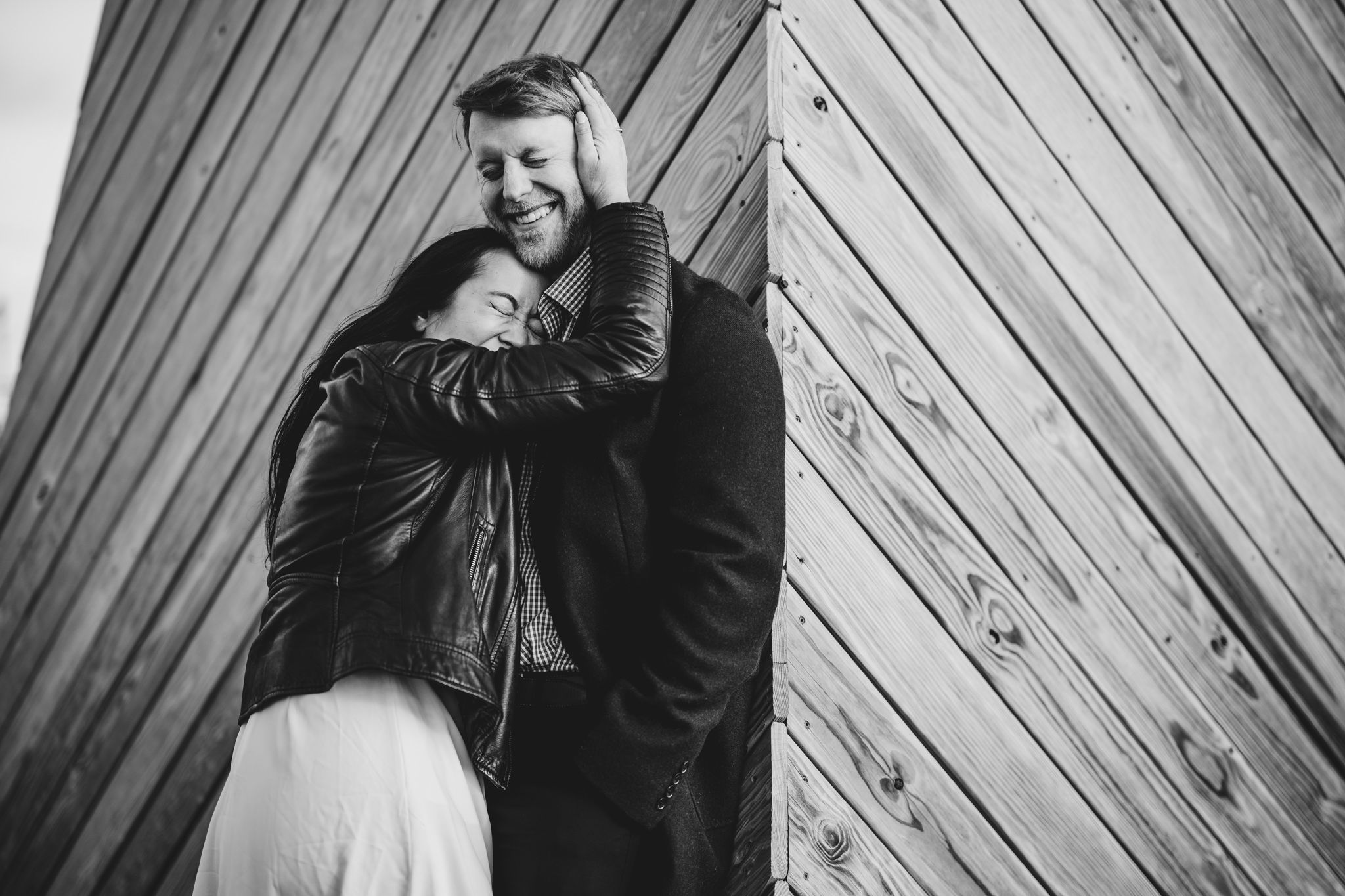 Black and white photo of a couple embracing and laughing against a diagonal wooden wall outdoors.