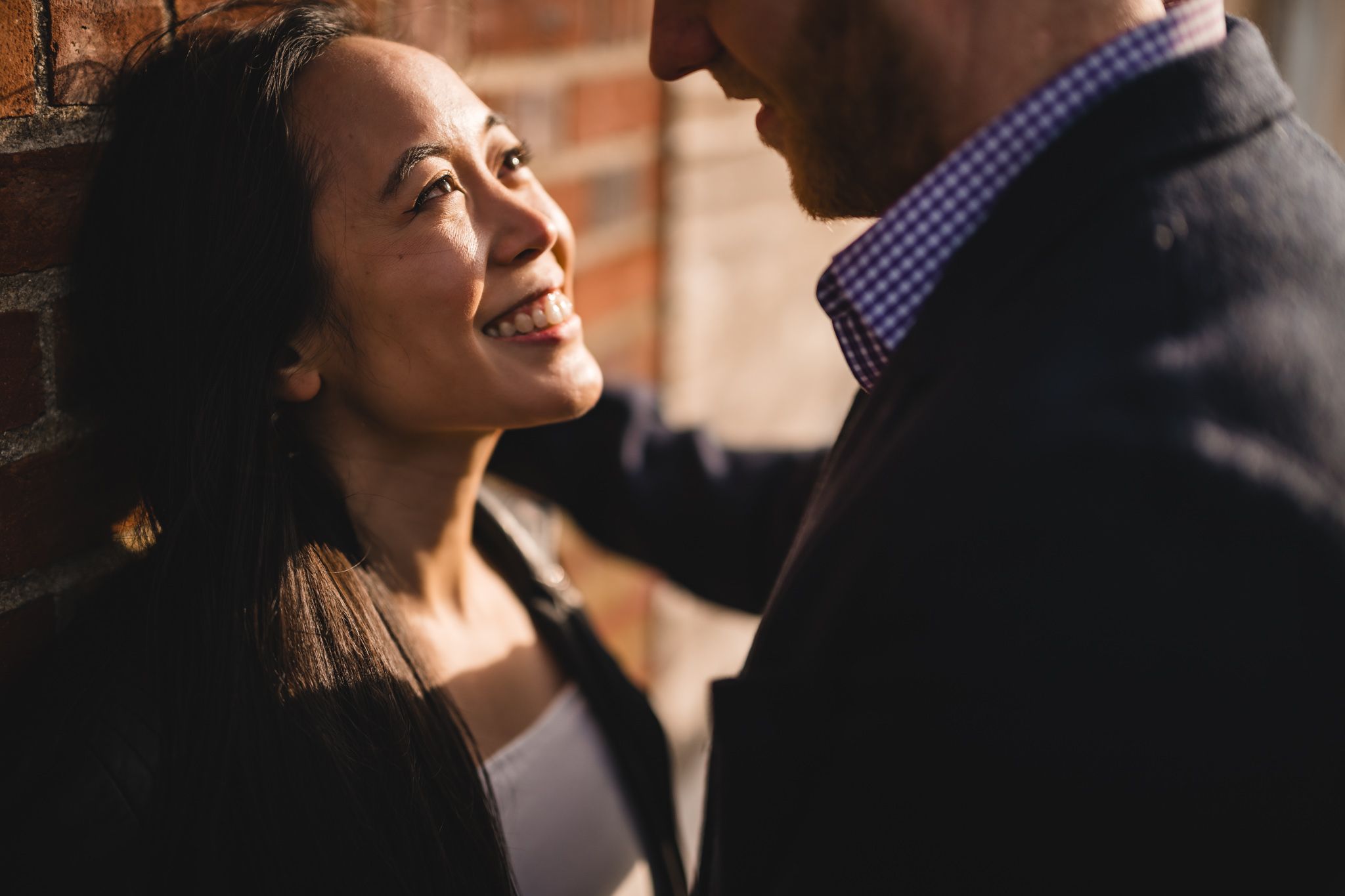 A smiling Asian woman leans against a brick wall while a man in a blazer faces her in a romantic moment.