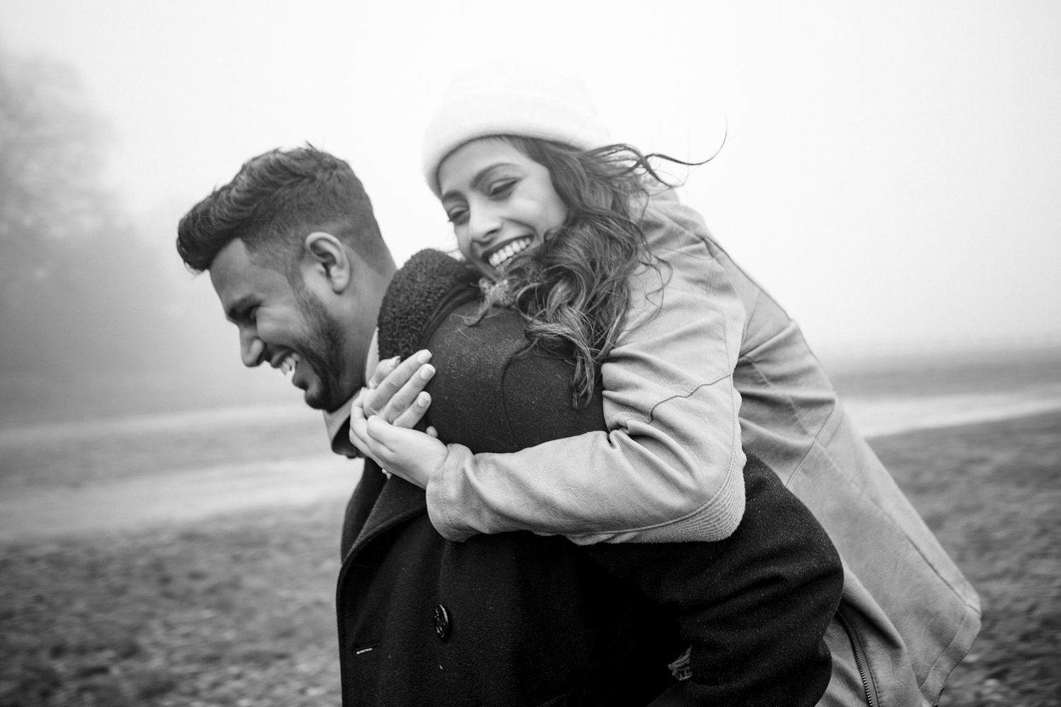 Black and white engagement photo of a joyful couple embracing outdoors on a foggy day, woman hugging man from behind.