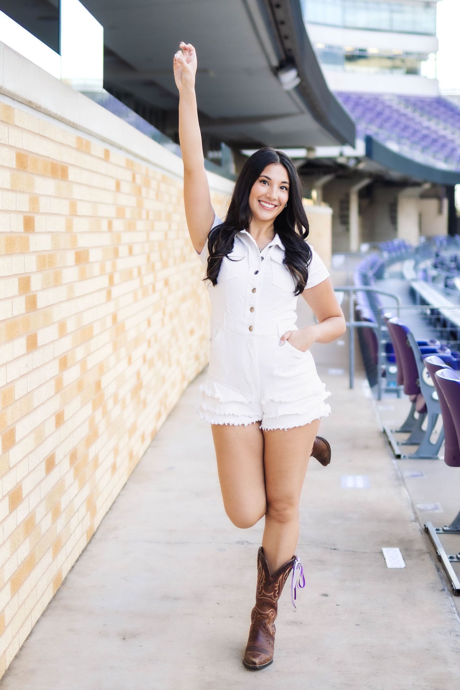 Young woman in white romper and cowboy boots poses cheerfully in a stadium concourse with arm raised.