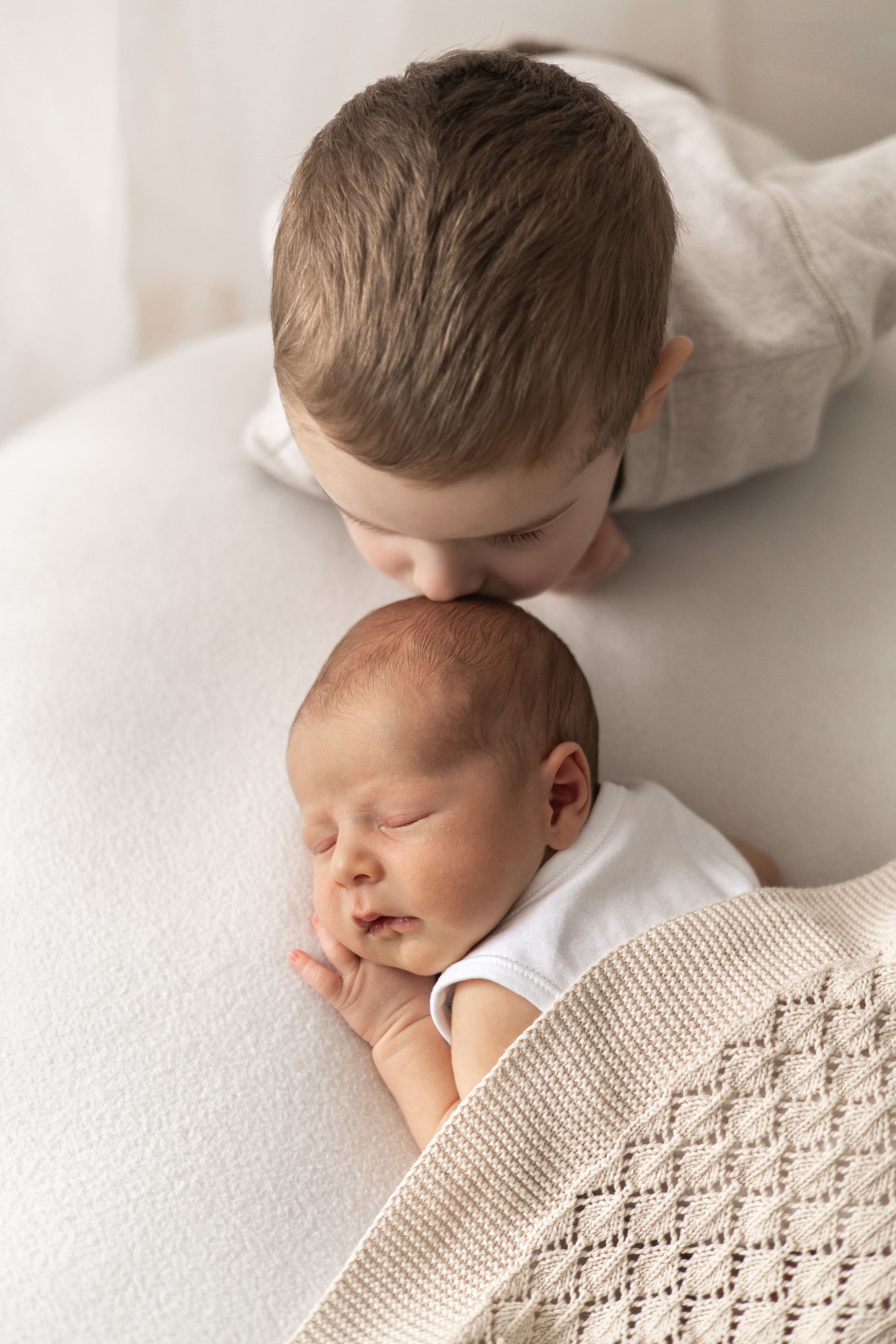 Older boy gently kissing sleeping newborn baby sibling wrapped in a cozy knit blanket Natural newborn photoshoot Nottingham