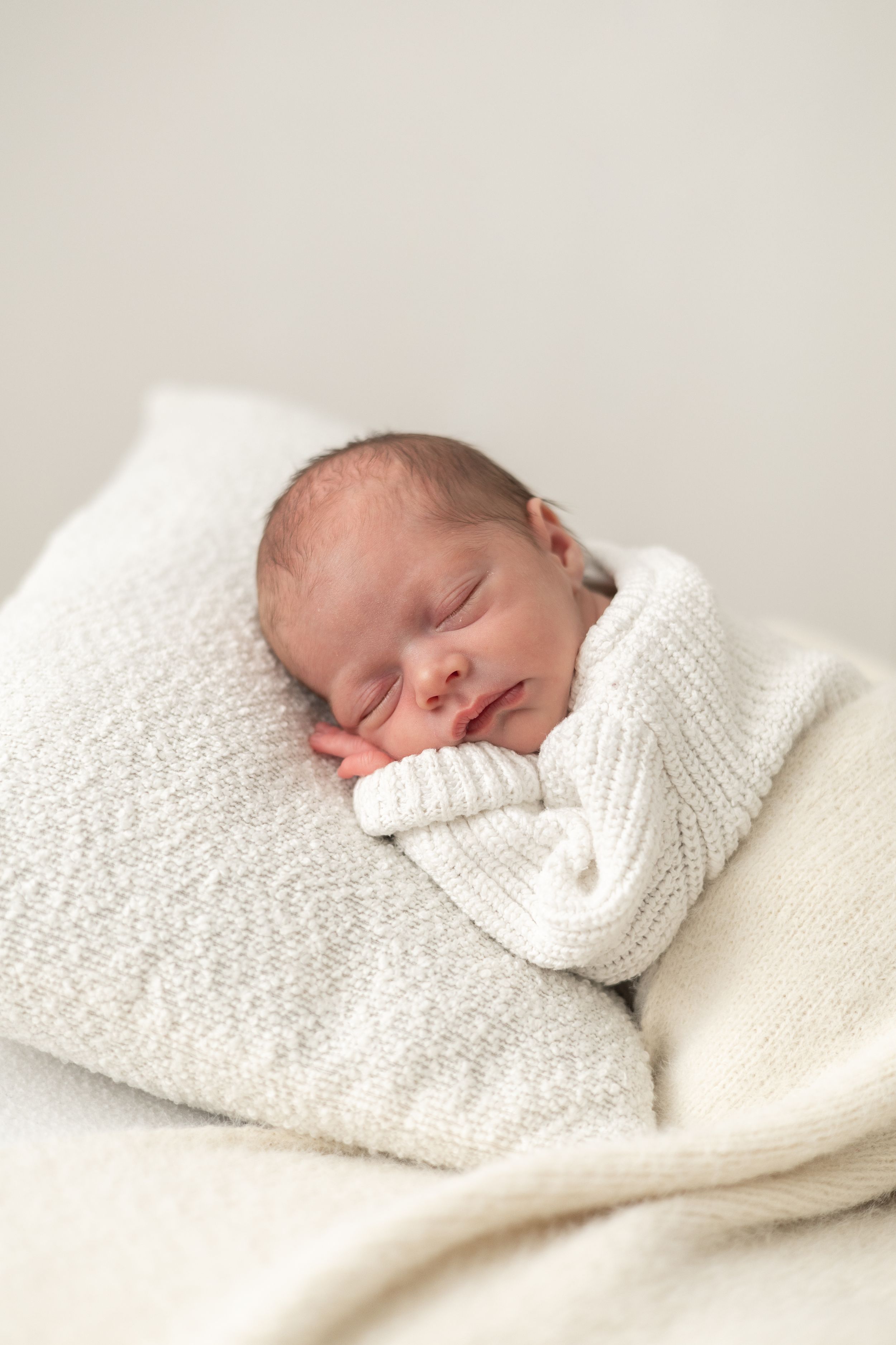 Peaceful newborn baby sleeping wrapped in a soft white knit blanket on a white pillow Natural newborn photoshoot Nottingham
