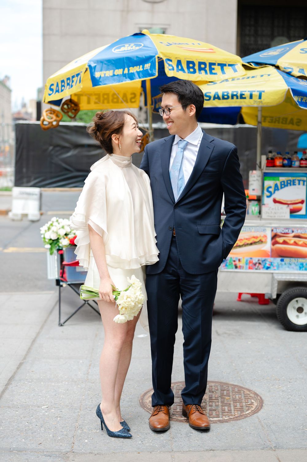 Couple posing by a Sabrett hot dog cart on a New York City street, bride holding white flowers.