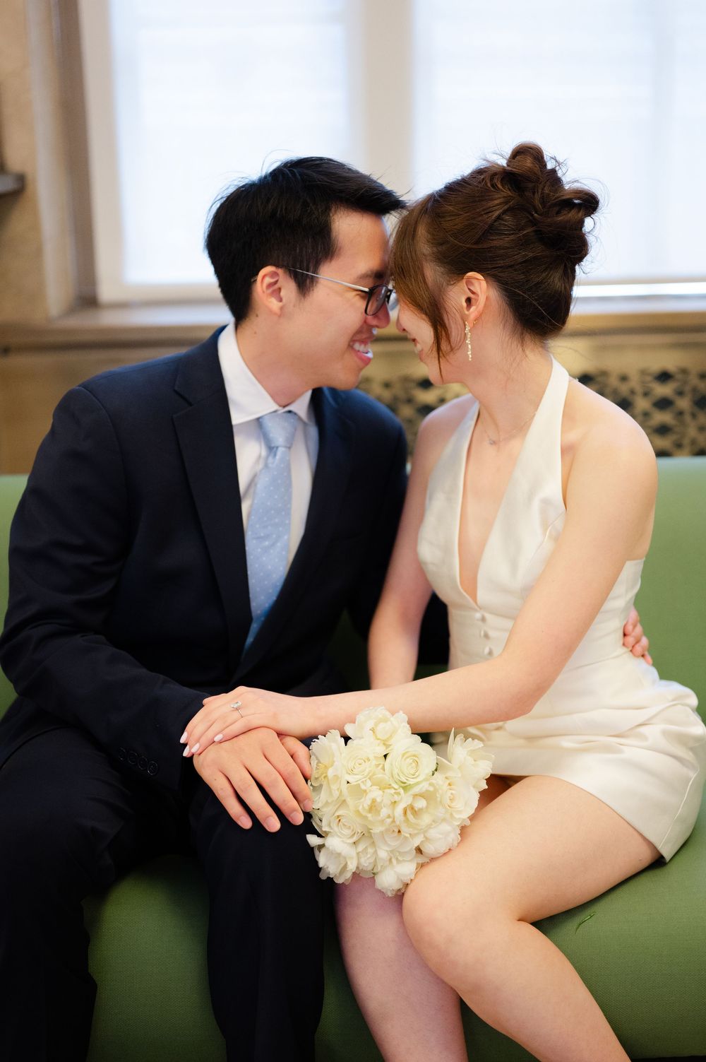Couple sharing an intimate moment on a green sofa, bride holding white peony bouquet.