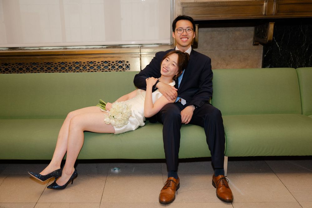 A couple in wedding attire poses on a green sofa, bride in white dress holding flowers, groom in dark suit.