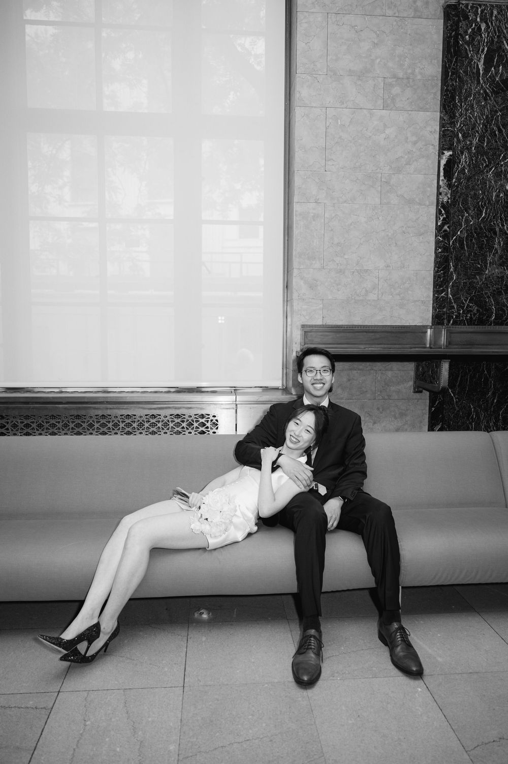 Black and white photo of bride leaning on groom while sitting on a bench in an elegant building lobby.