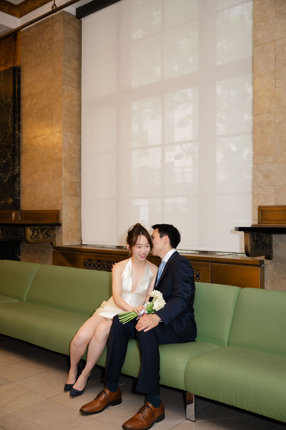 Bride and groom sitting closely on a green bench in a marble-walled building lobby sharing an intimate moment.