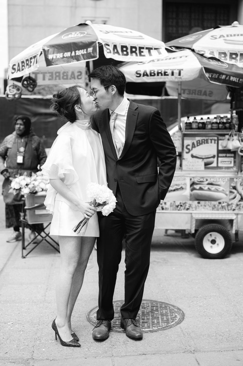 Couple sharing a kiss in front of a Sabrett hot dog cart on a New York City street in wedding attire.