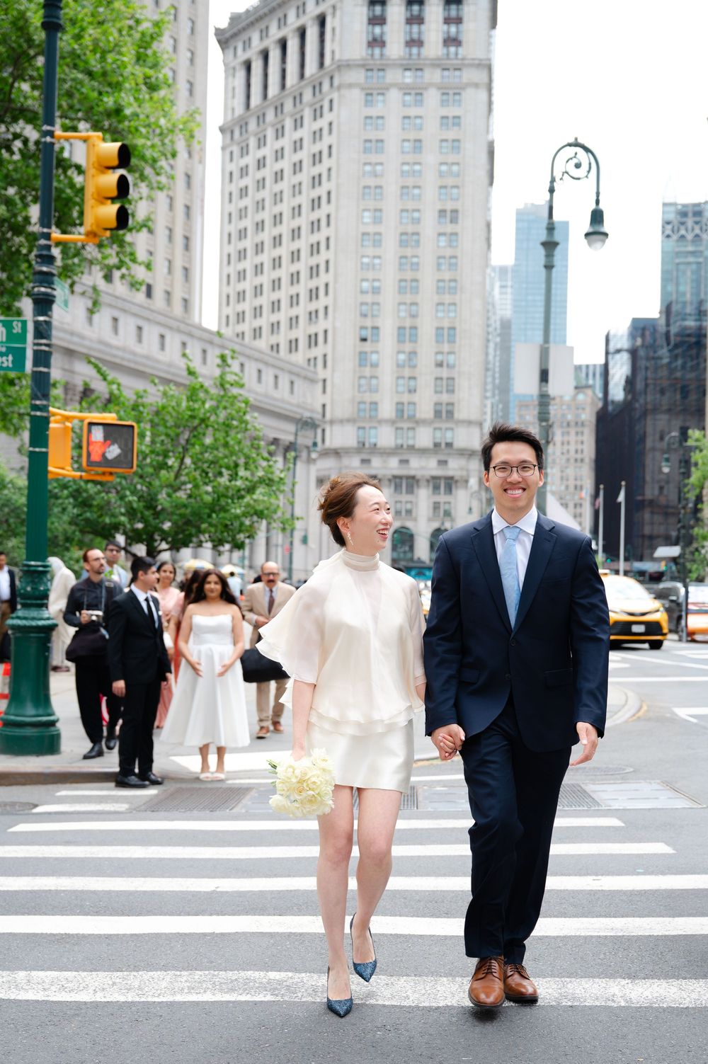 Newlyweds holding hands crossing a Manhattan street, bride in white mini dress with bouquet.