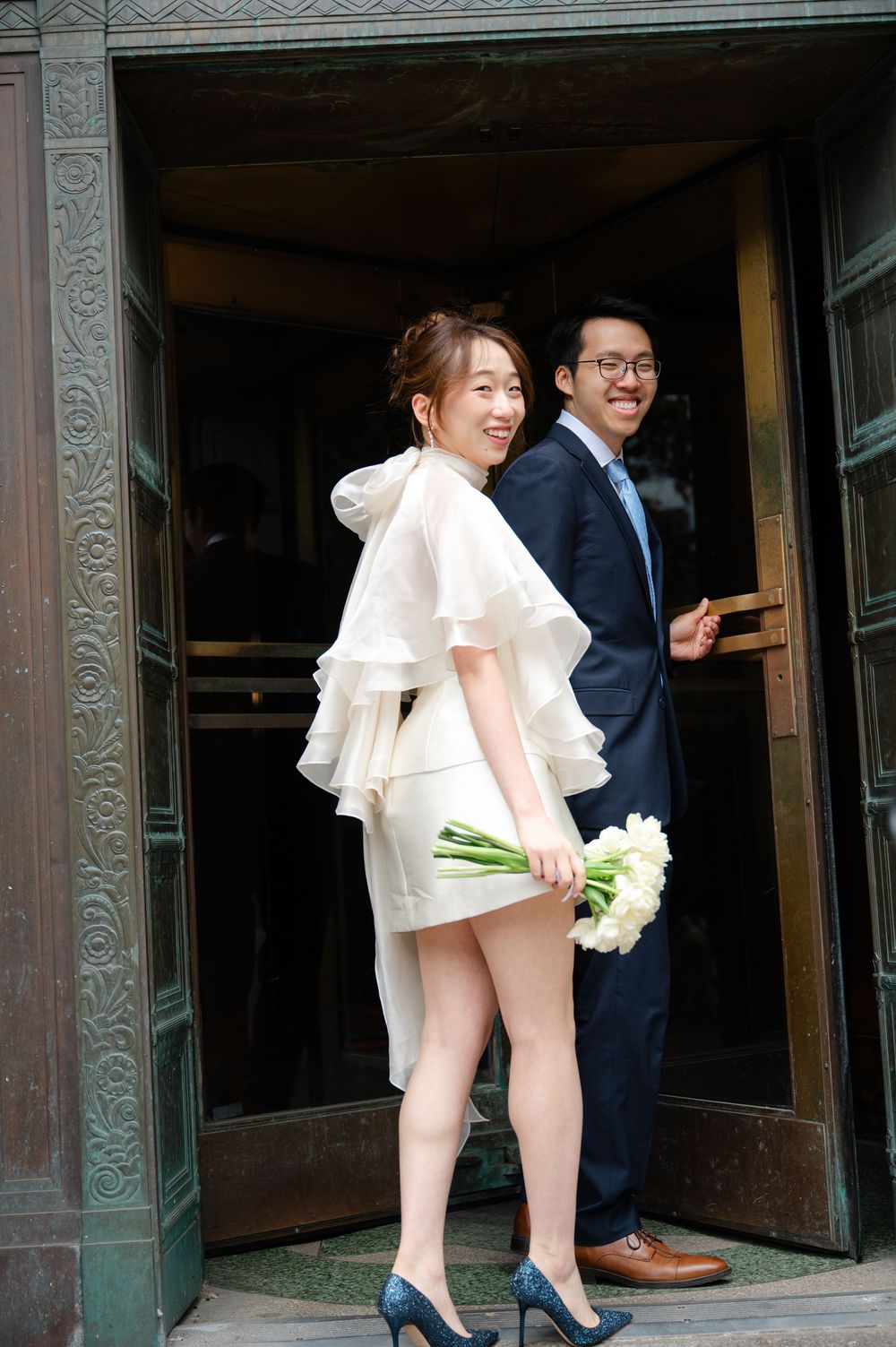 Bride in ruffled white mini dress and groom in navy suit posing at ornate building entrance holding tulips.