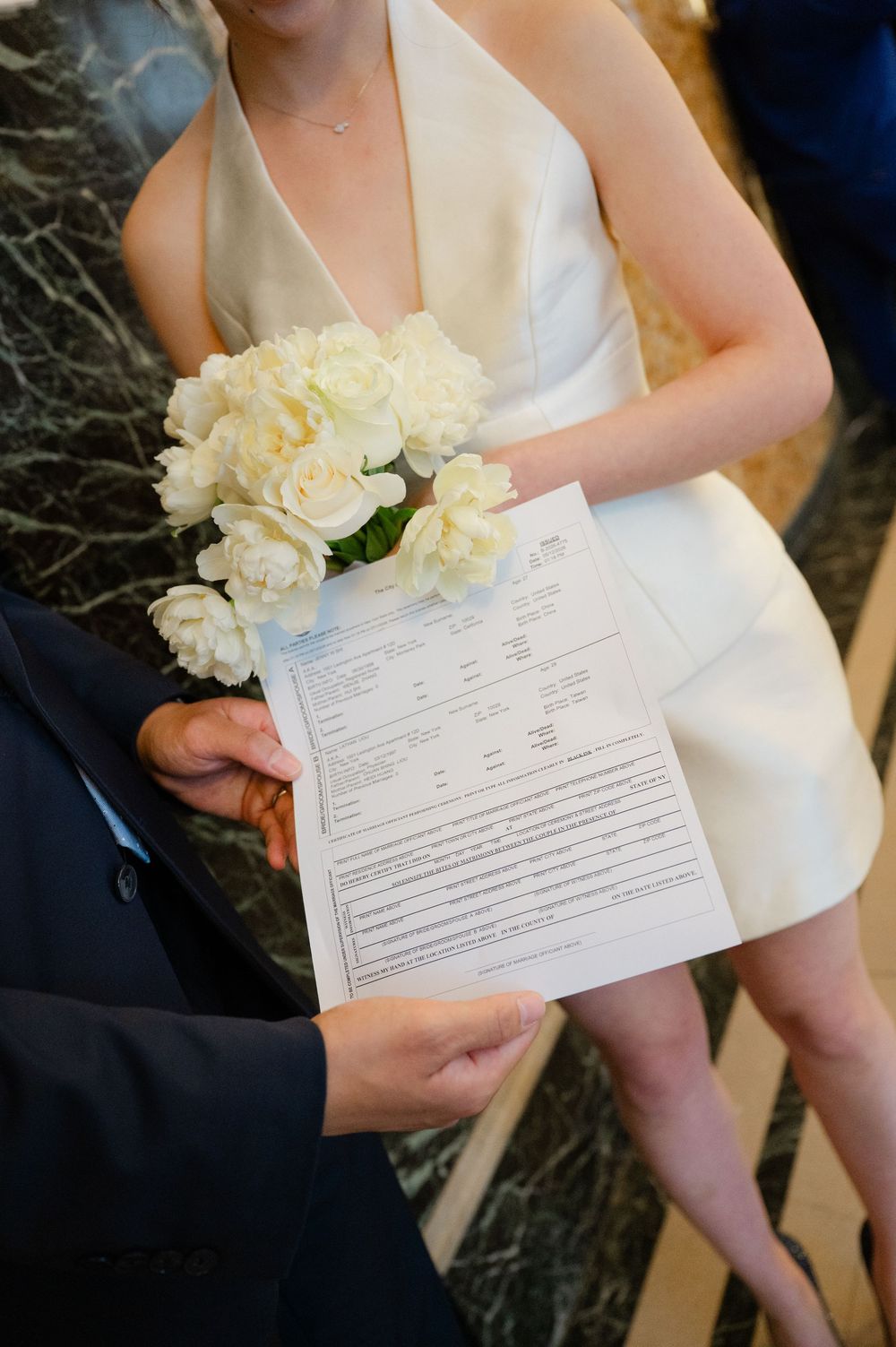 Bride in white mini dress holds bouquet while couple reviews marriage license document.