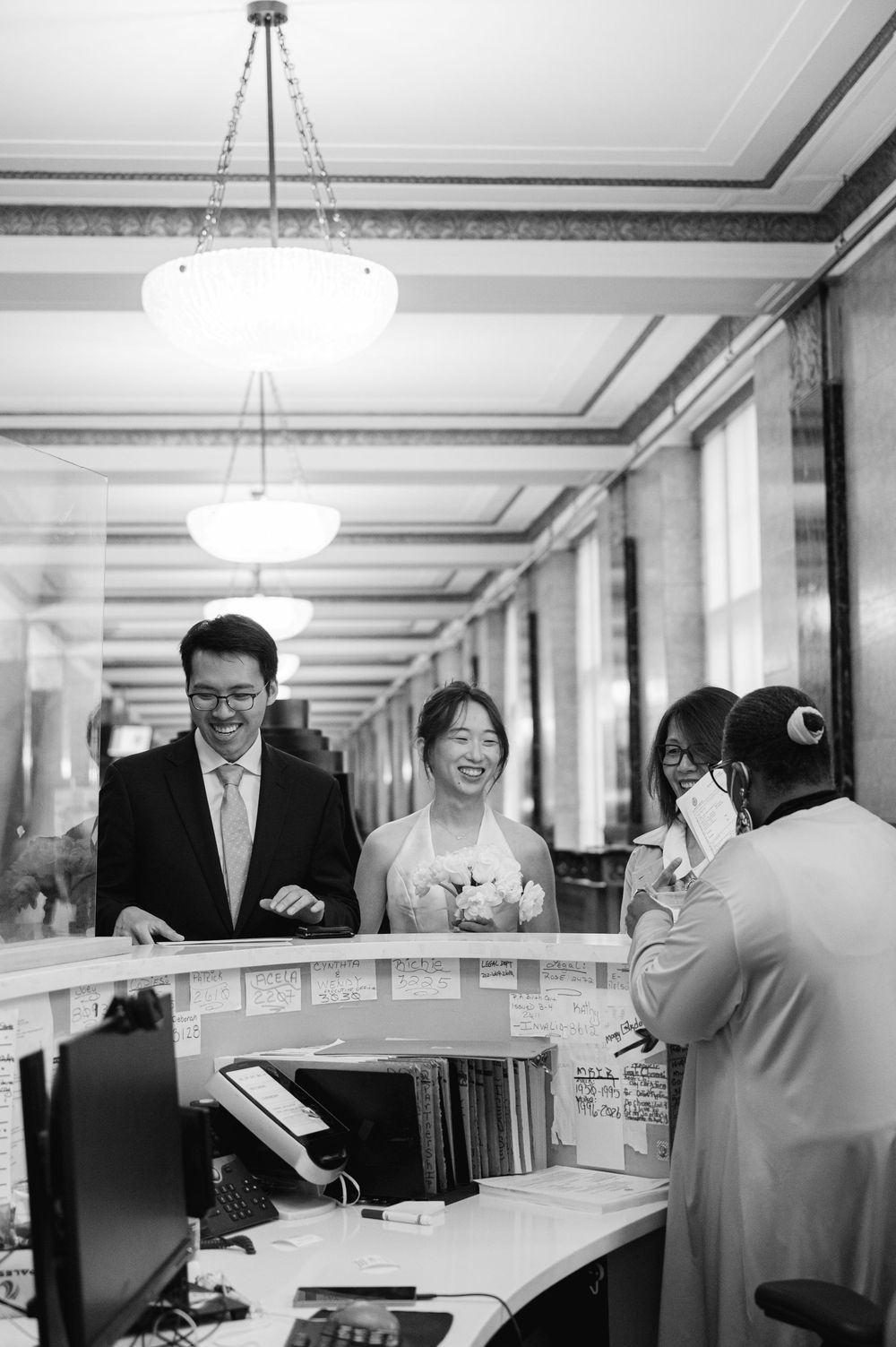 Bride and groom smiling at NYC City Hall clerk during marriage registration in black and white photo.