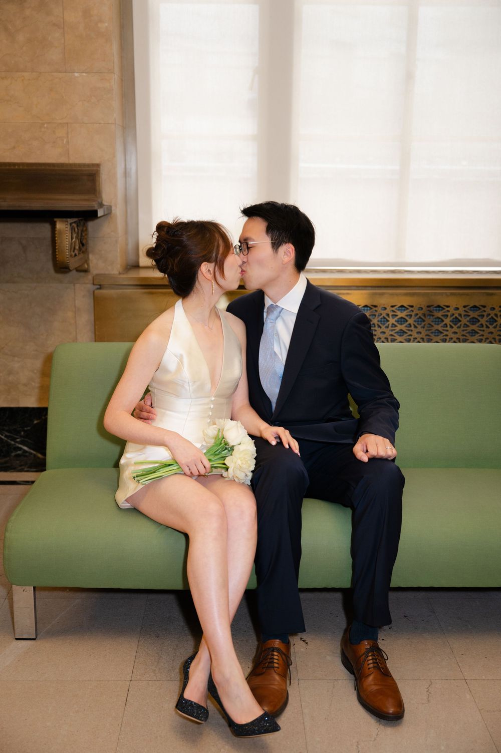 Bride and groom share a kiss while seated on a green sofa, bride holding white bouquet in elegant short wedding dress.