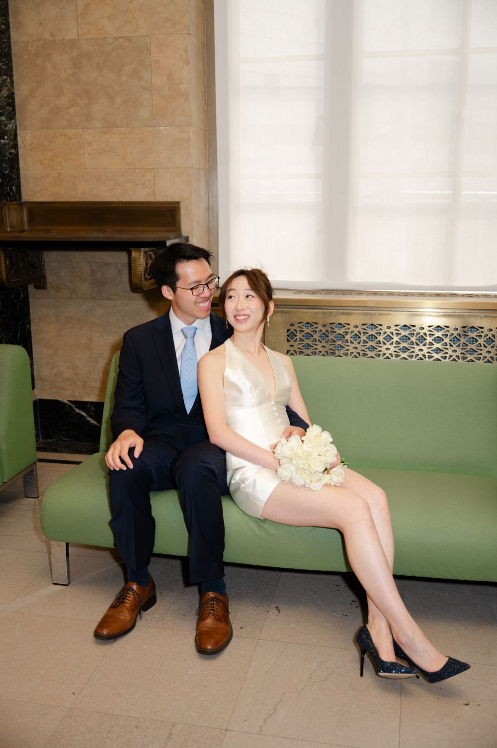 Couple in wedding attire sitting on green sofa, bride in white mini dress holding bouquet, groom in navy suit.