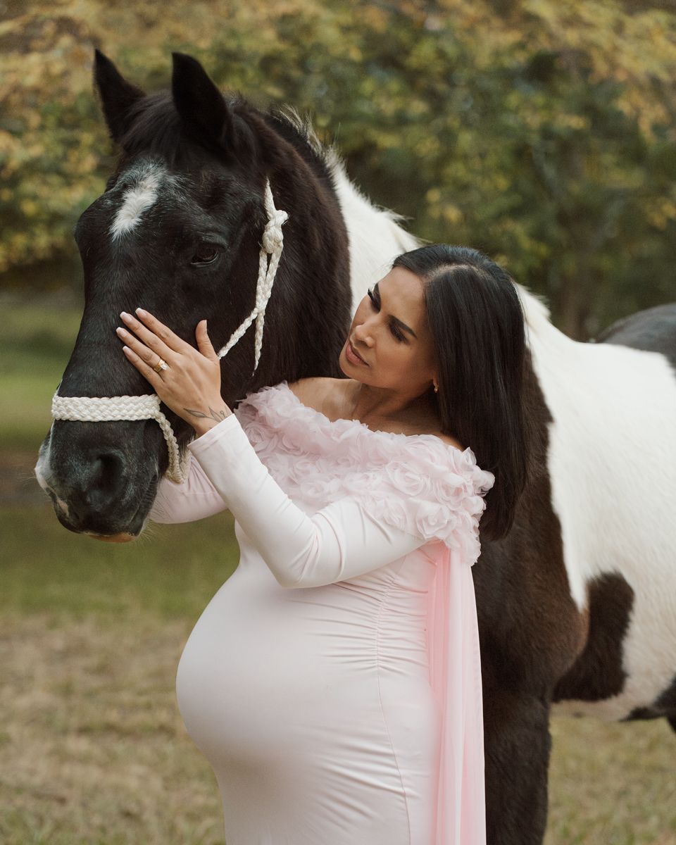 Pregnant woman in pink dress tenderly touches black and white paint horse outdoors in autumn setting photoshoot with a horse in sydney