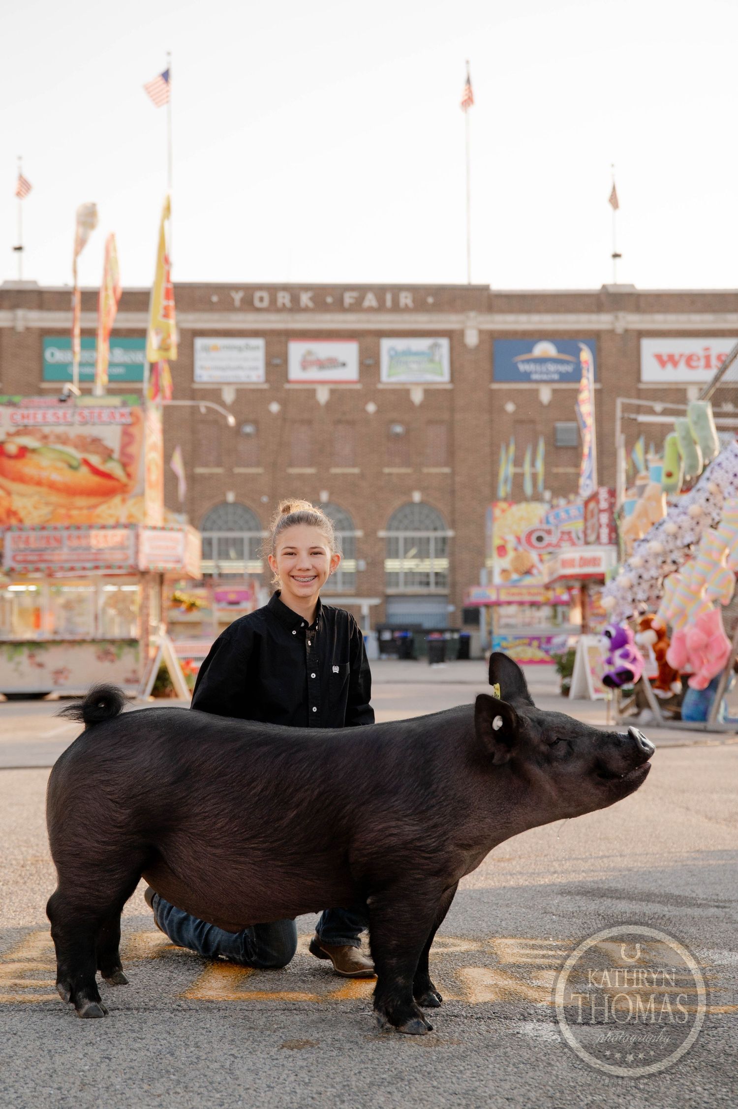 Young woman smiling beside a large black pig at York Fair, with carnival rides and fair entrance visible in background.