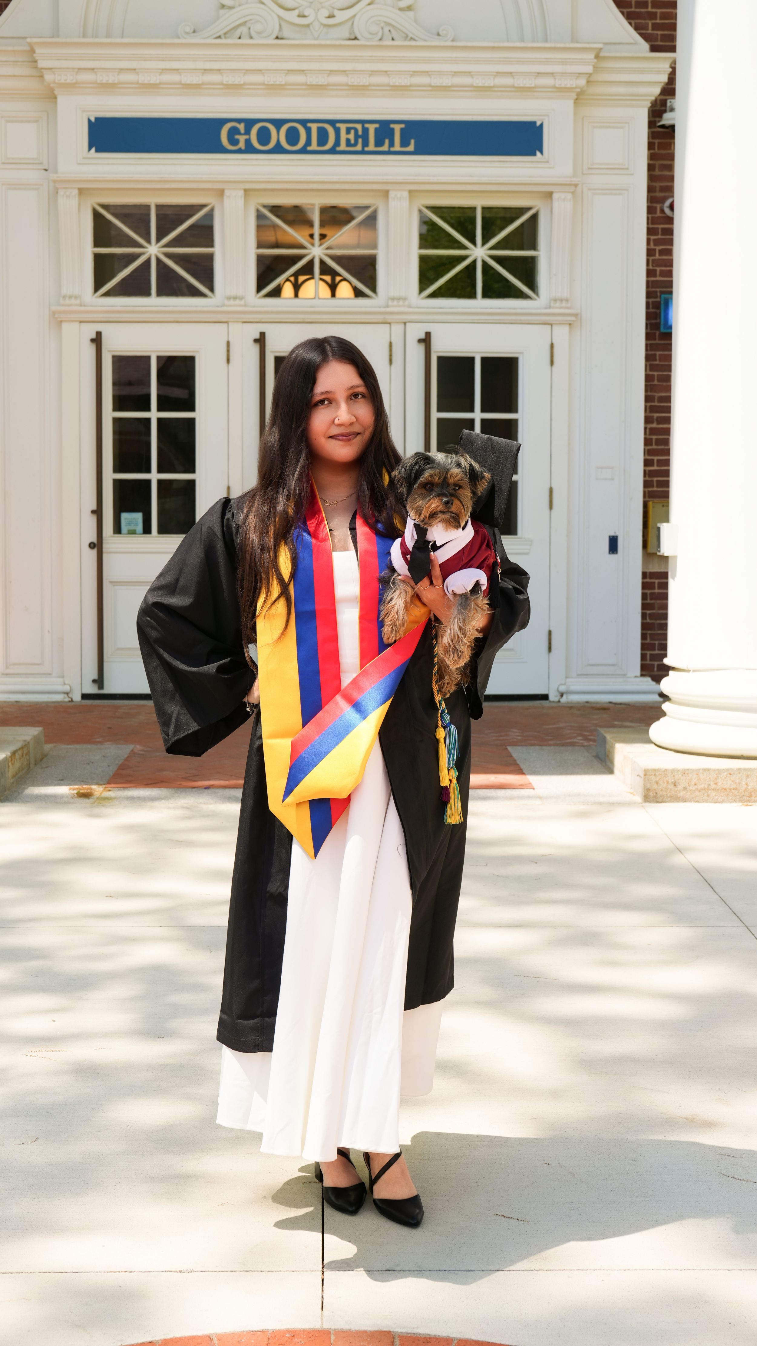 Graduate in black gown with colorful sash holds small dog outside Goodell building on graduation day.