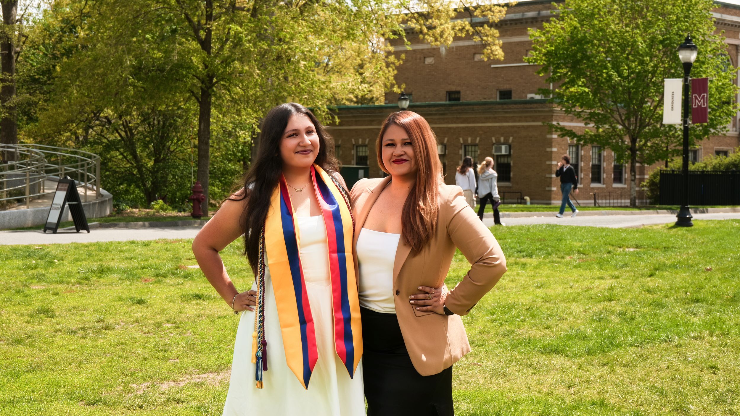Two women posing outdoors at a graduation ceremony, one wearing colorful academic sashes on a sunny campus.