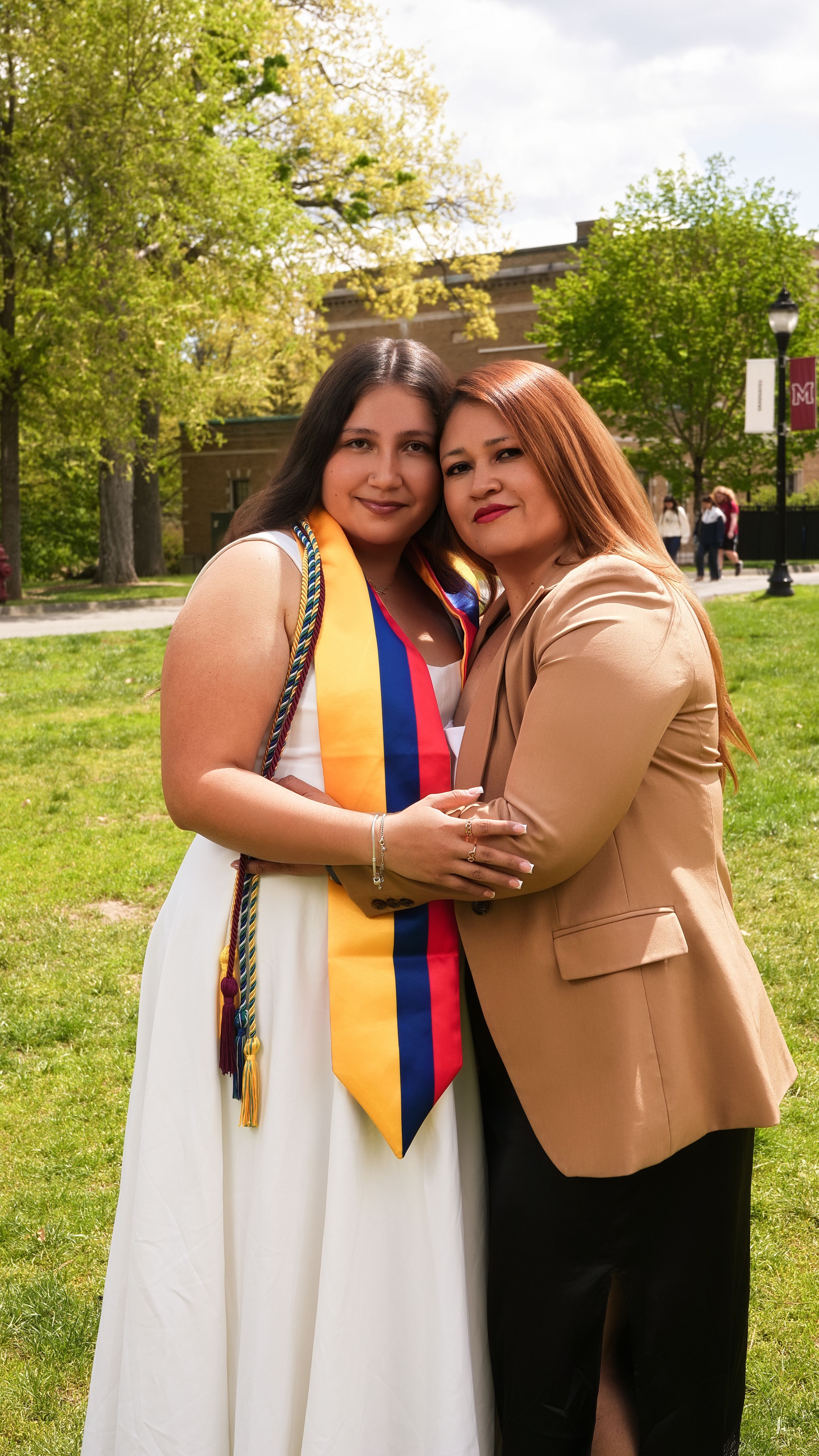 Two women embrace outdoors at a graduation ceremony, one wearing a white dress with colorful academic stoles.