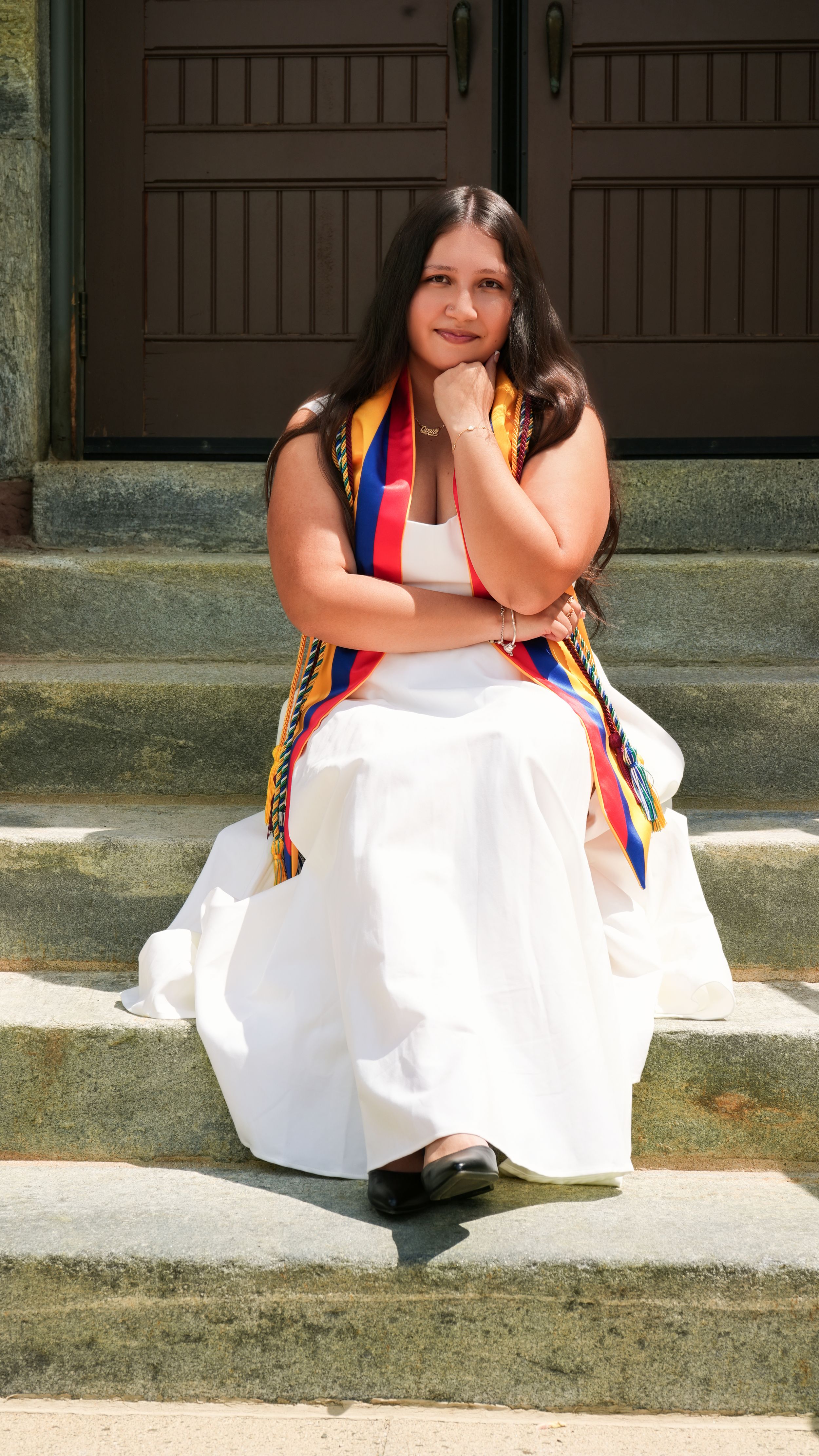 Young woman in white dress and colorful graduation stole sits on stone steps, posing for graduation photo.